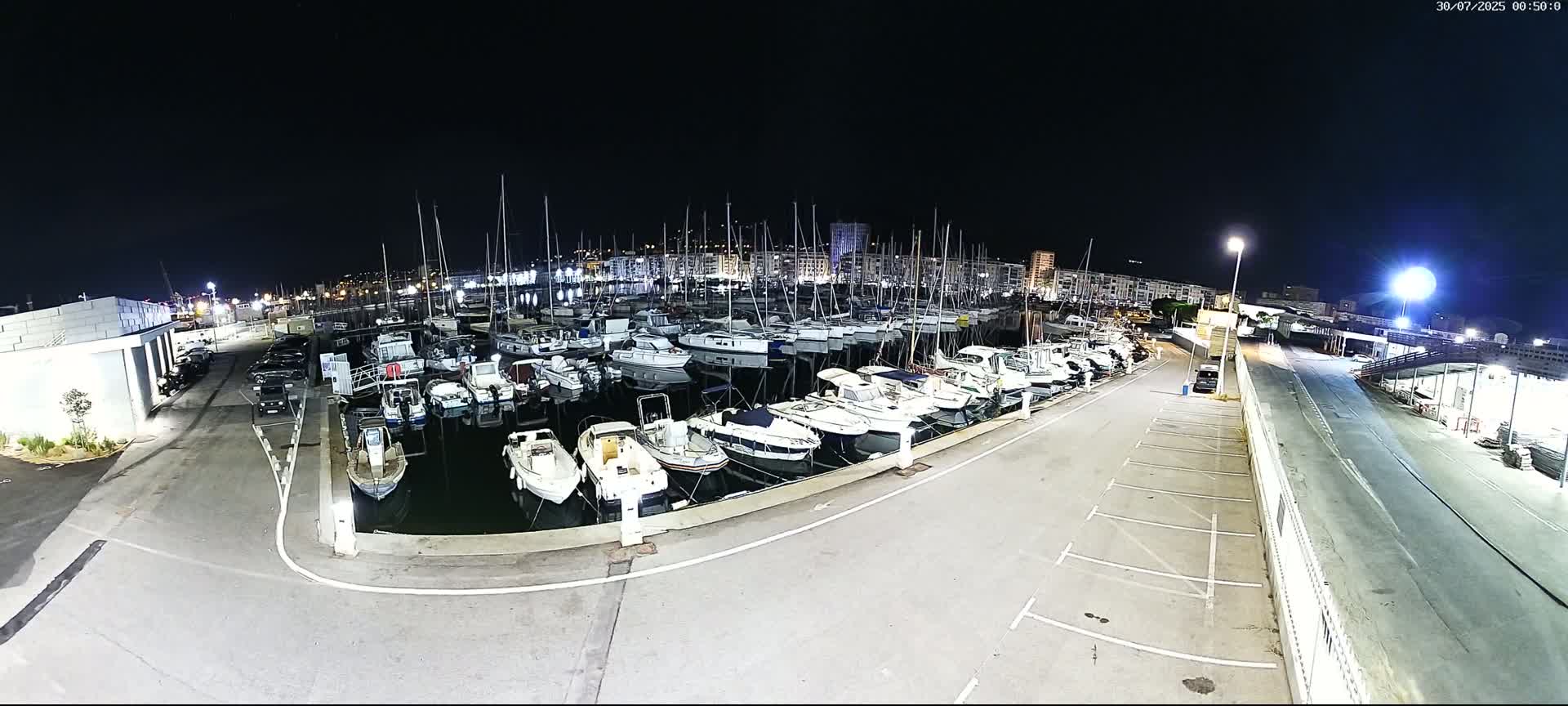 A marina filled with numerous boats is situated near a city under a cloudy sky, with a road and parking lot adjacent to the waterfront.