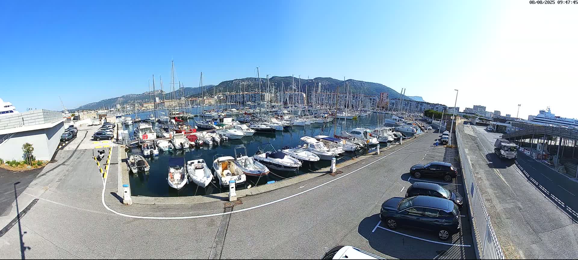 A marina filled with numerous boats is seen under a clear blue sky, with cars parked along a roadway adjacent to the water and mountains visible in the background.
