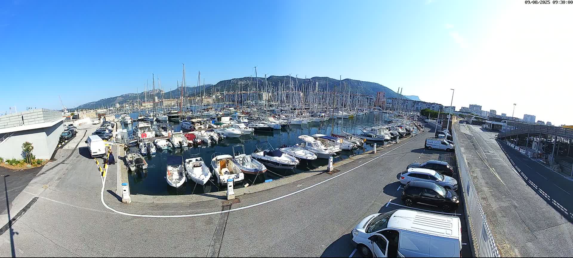 A marina filled with numerous boats of various sizes is shown under a clear, sunny sky, with a parking lot and distant buildings visible.