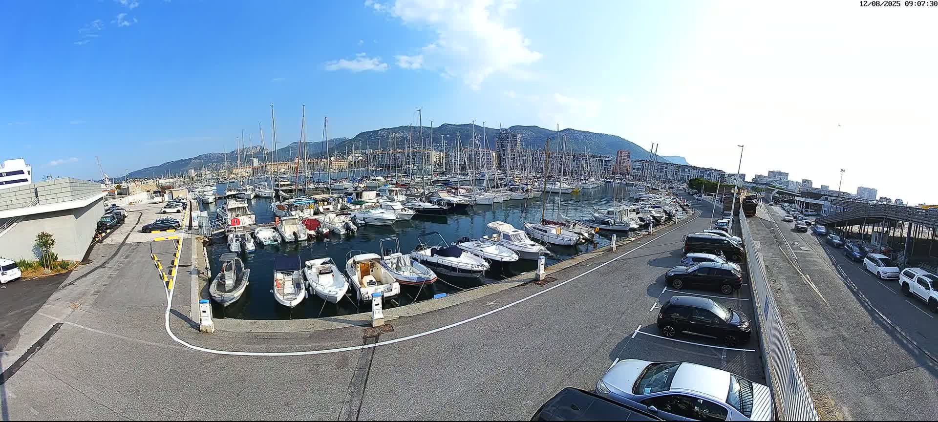 A marina filled with numerous boats is shown under a sunny sky, with a parking lot and road visible nearby, and mountains in the distance.