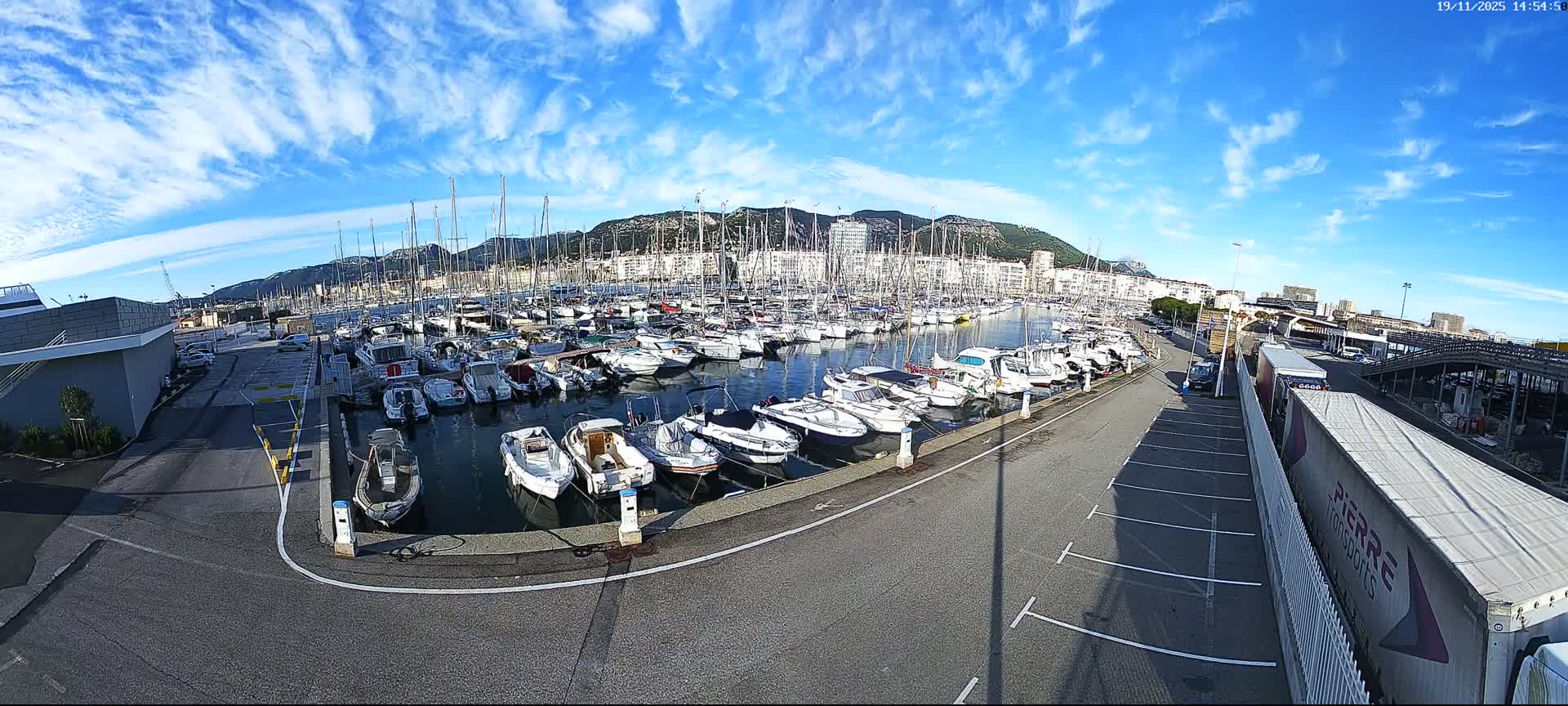 A marina filled with numerous boats is shown under a sunny sky, with a parking lot and road visible nearby, and mountains in the distance.