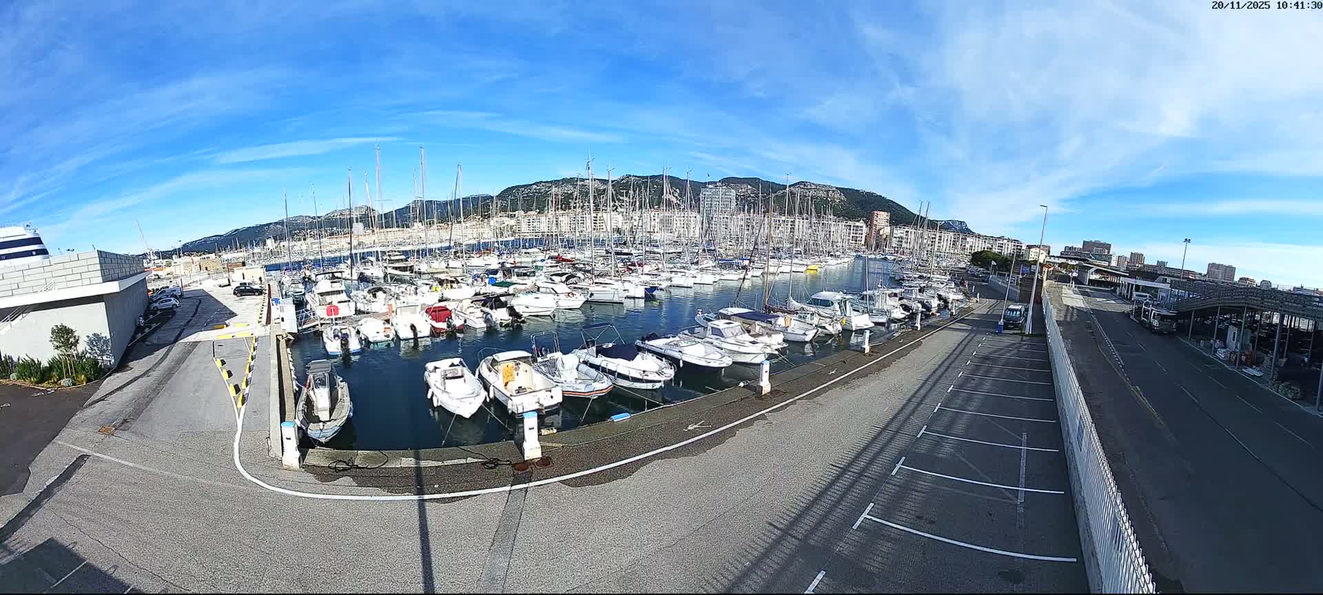 A wide-angle shot captures a bustling marina filled with numerous docked boats, surrounded by buildings and roads, all set against distant mountains under a bright, sunny blue sky with scattered wispy clouds.