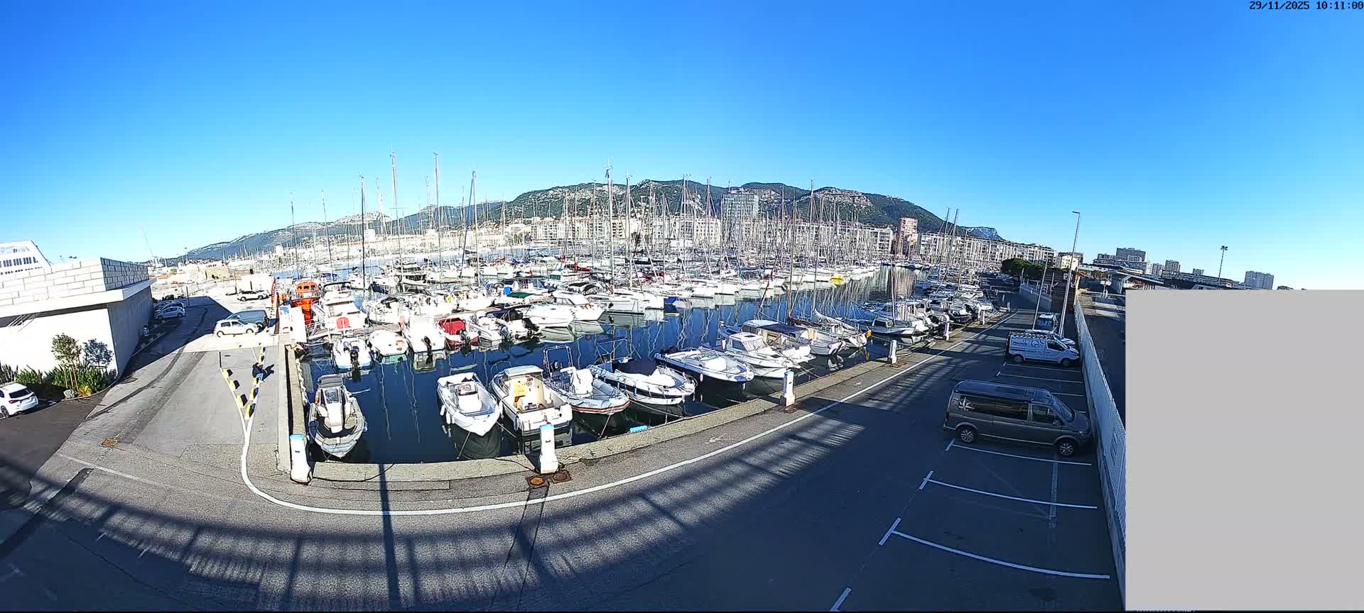 A bustling marina filled with numerous boats and yachts is seen from above on a sunny day with a clear blue sky, backed by a city and mountains.