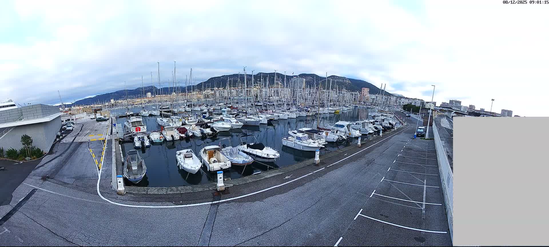 A wide-angle view captures a busy marina filled with numerous boats and sailboats docked along a paved waterfront with parking, all under an overcast sky and backed by distant mountains and urban buildings.