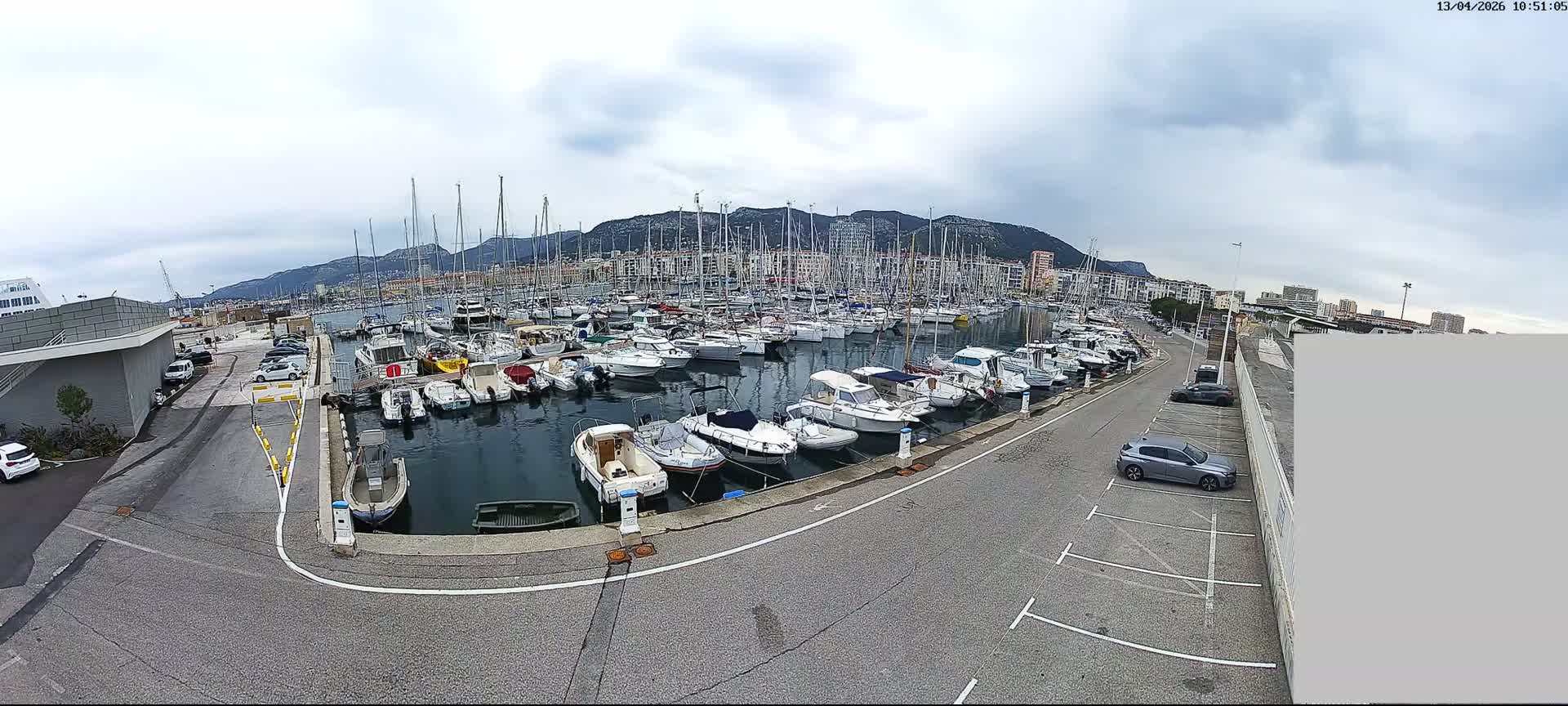 A wide-angle view captures a busy marina filled with numerous boats and sailboats docked along a paved waterfront with parking, all under an overcast sky and backed by distant mountains and urban buildings.