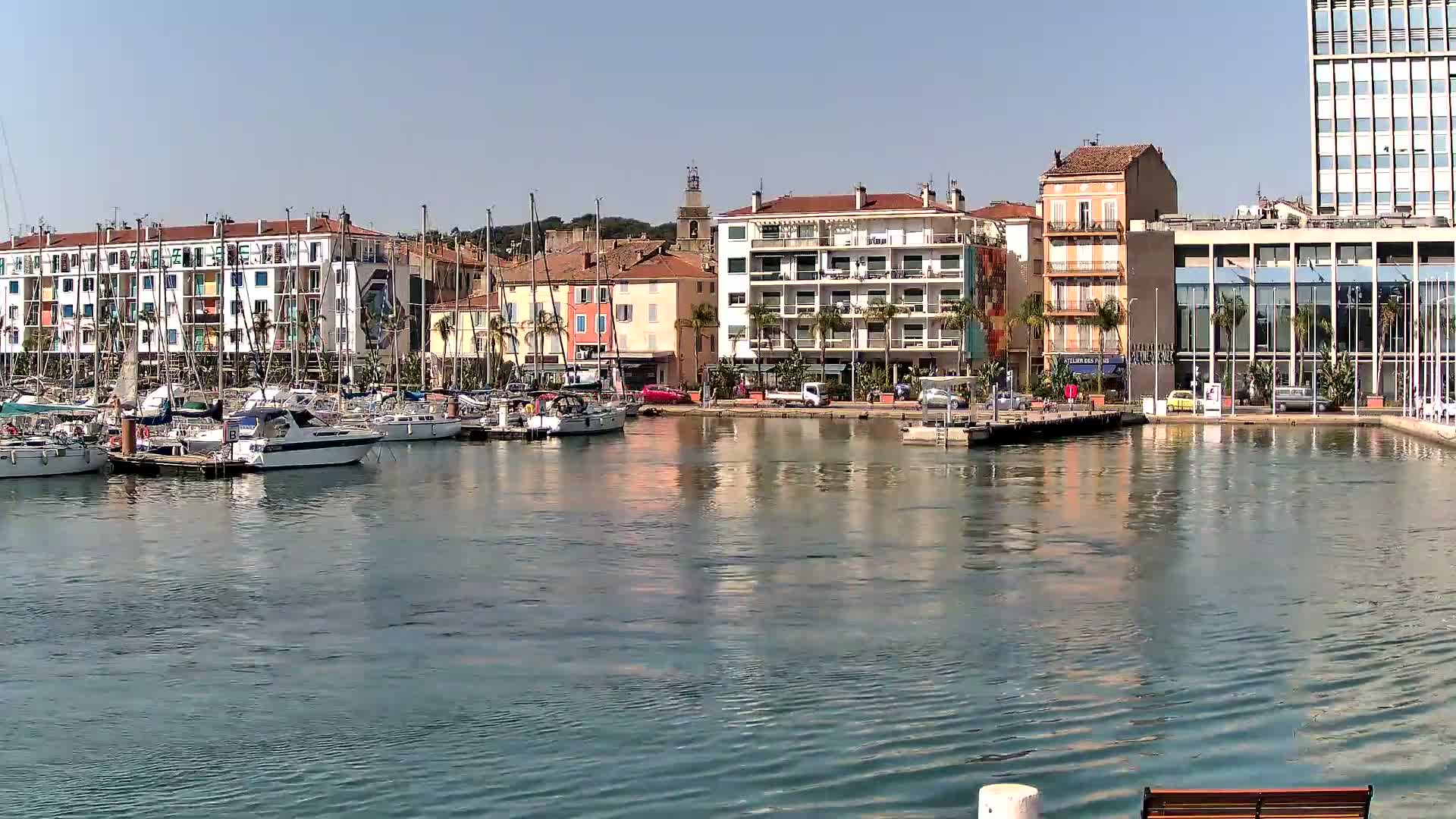 A calm harbor filled with sailboats and motorboats is nestled beside multi-story buildings under a clear blue sky.