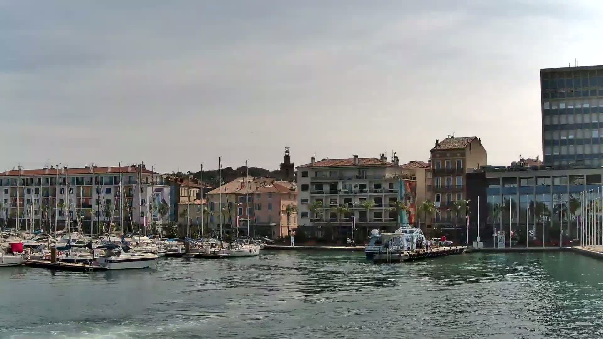 A calm harbor filled with sailboats and motorboats is nestled beside multi-story buildings under a clear blue sky.