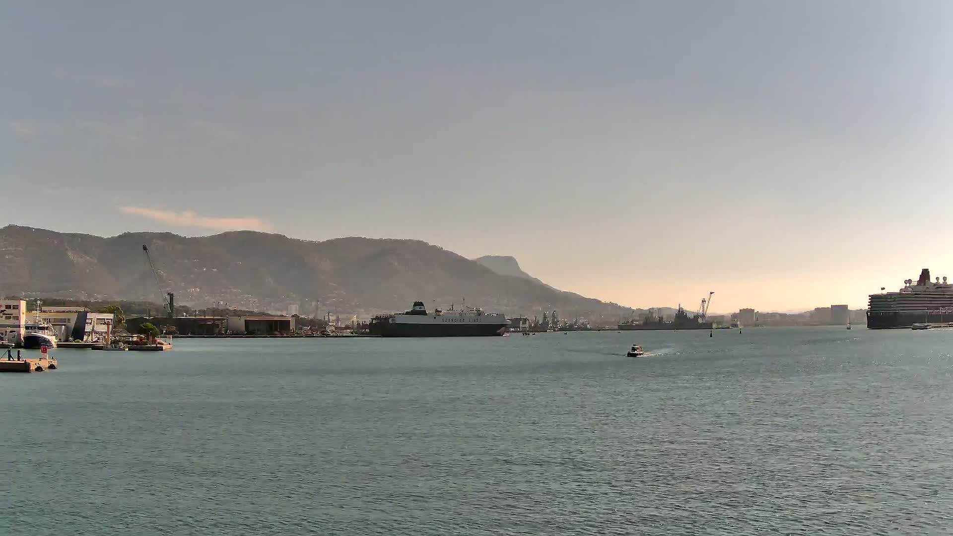 A calm harbor scene shows several ships, including a large cargo ship and a cruise ship, with mountains in the background under a clear sky.
