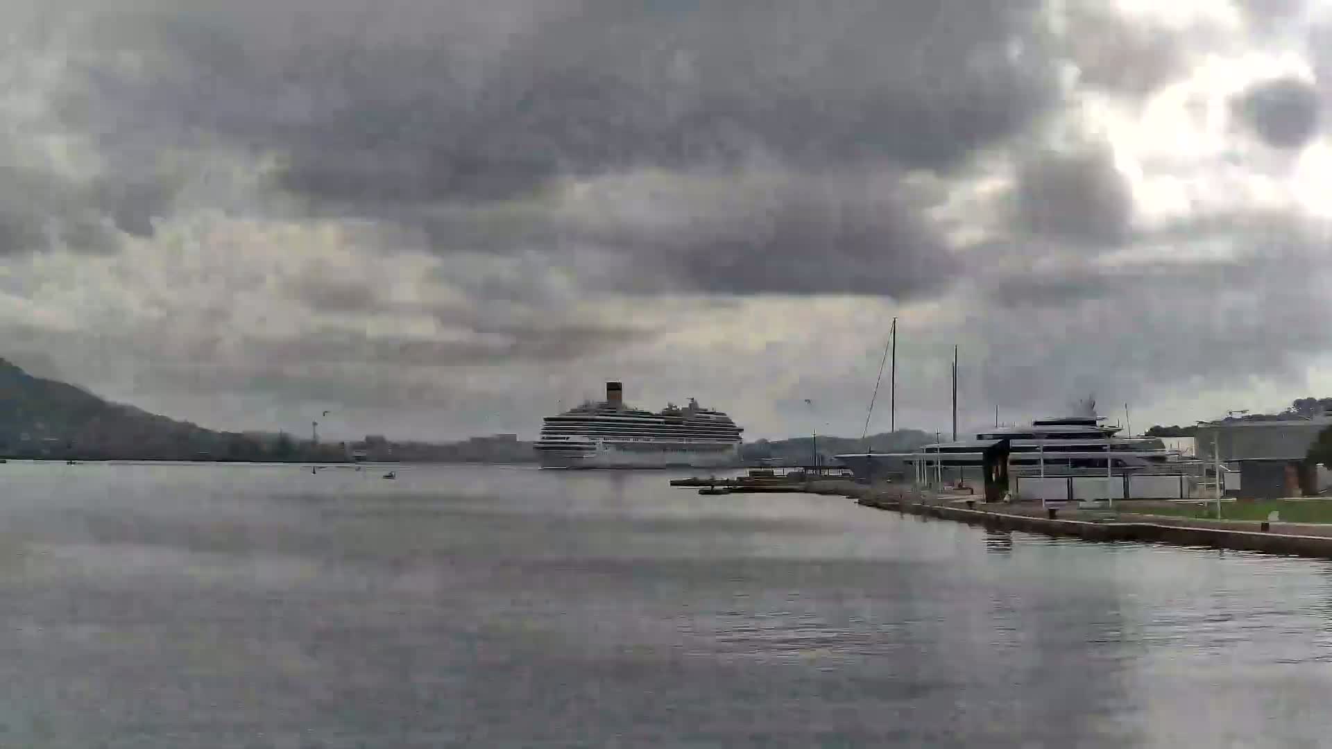 A large cruise ship and a smaller yacht are docked in calm water under a cloudy sky.