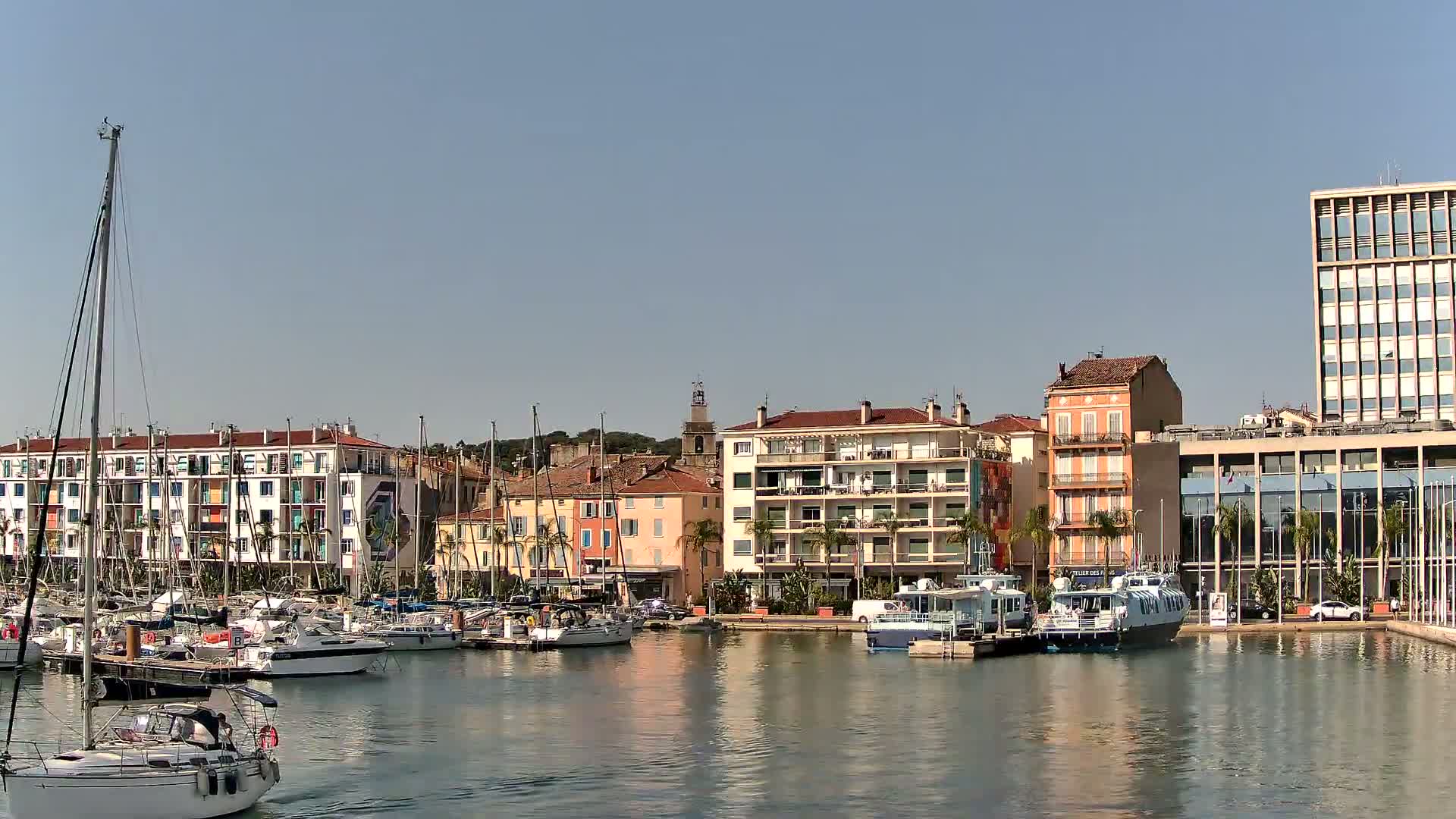 A marina filled with sailboats and motorboats is situated in front of a waterfront town featuring multistory buildings under a clear, sunny sky.