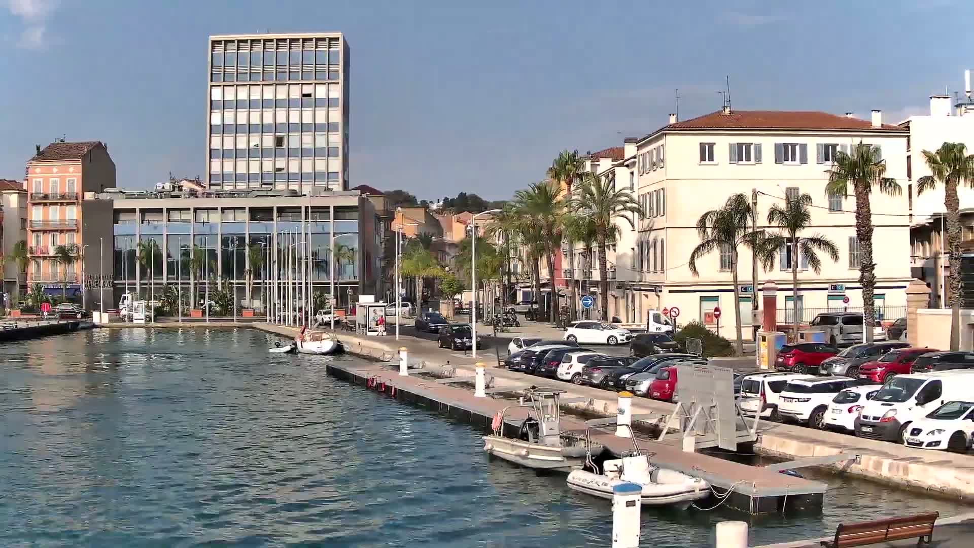 A marina scene on a sunny day shows several boats docked alongside a waterfront lined with buildings, palm trees, and parked cars.