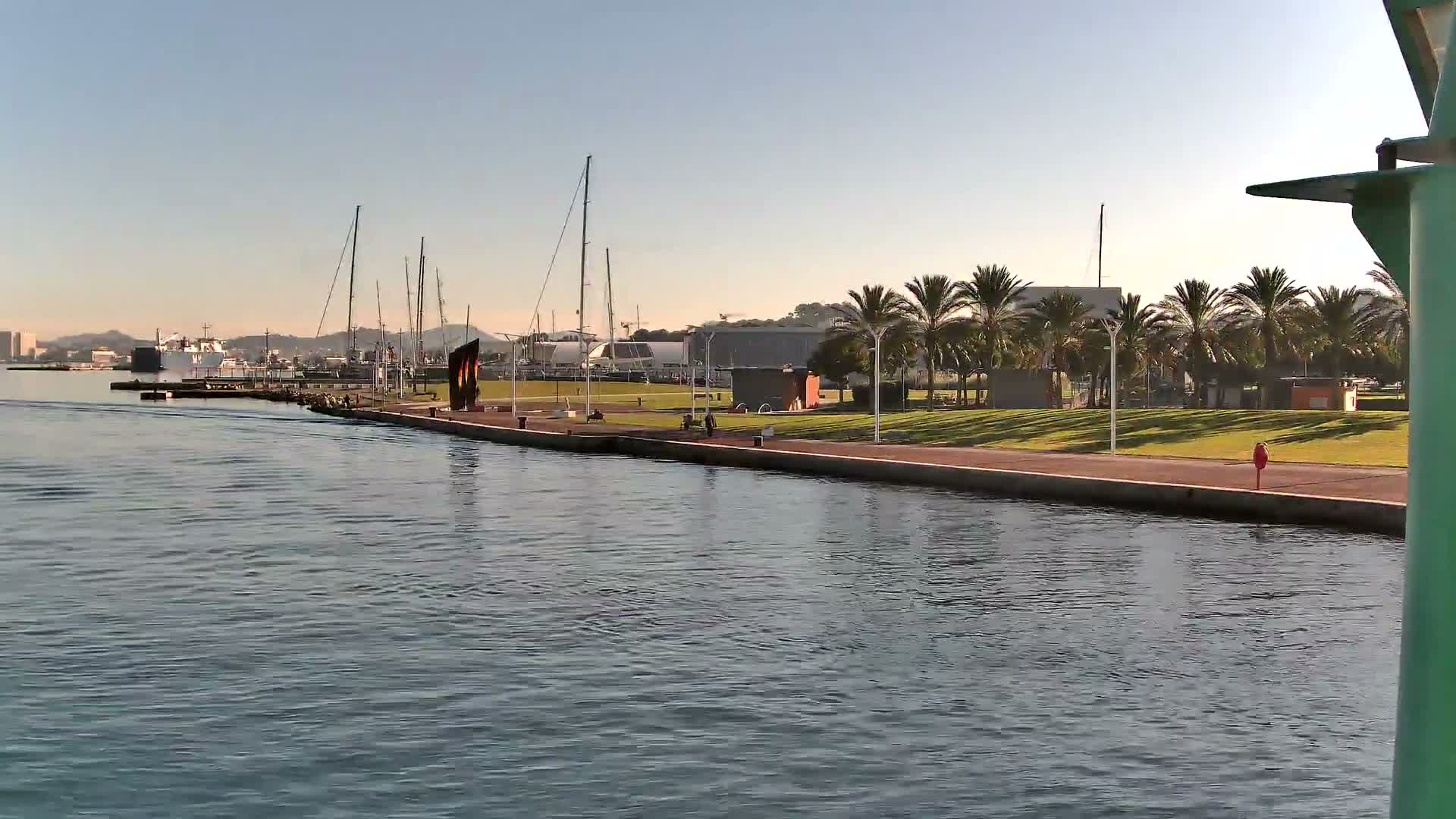 A sunny harbor scene features calm blue water, a promenade lined with palm trees and a few pedestrians, several moored sailboats, and distant hills under a clear, bright sky.