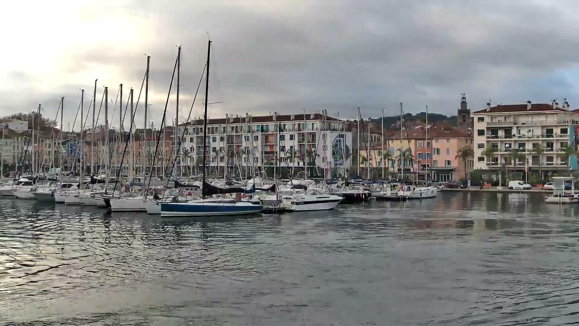 Numerous sailboats and motorboats are moored in a lively marina, framed by a row of diverse buildings and a distant church tower, all beneath a uniform overcast sky.