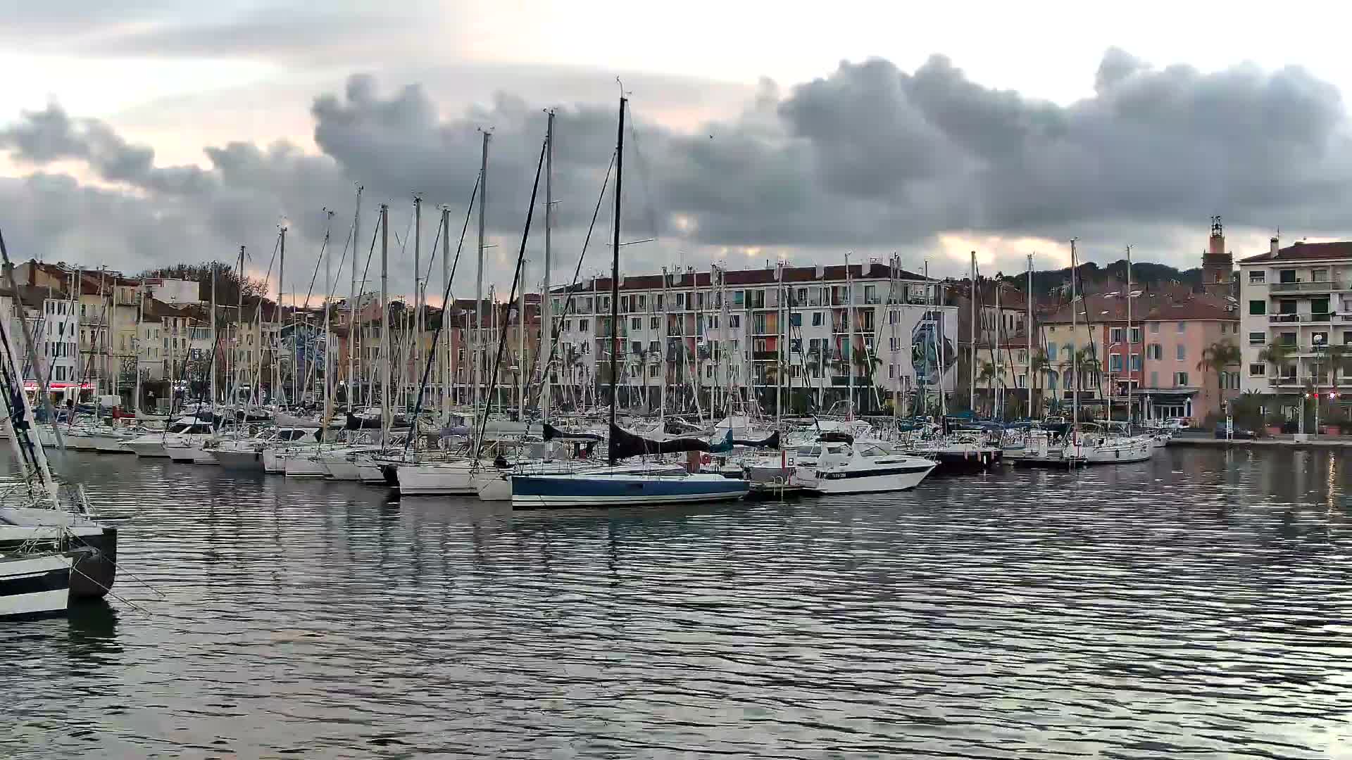 Numerous sailboats and motorboats are moored in a lively marina, framed by a row of diverse buildings and a distant church tower, all beneath a uniform overcast sky.