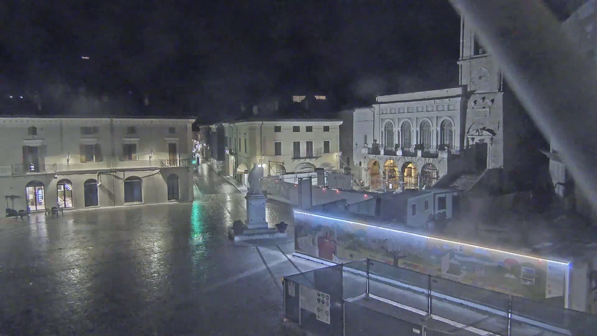 A rain-slicked town square at night, featuring several buildings, a statue, and construction equipment.
