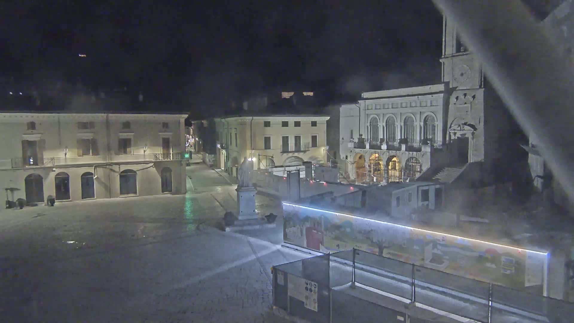 A nighttime view of a town square shows several buildings, a statue, and construction barriers in the center of the square.