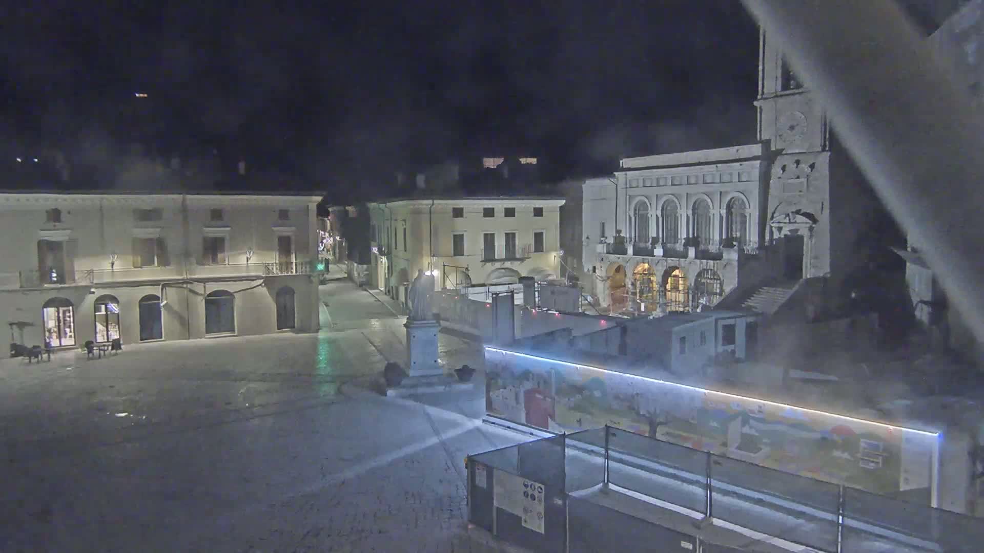 A snow-covered town square at night, featuring several buildings, a statue, and construction barriers with artwork.