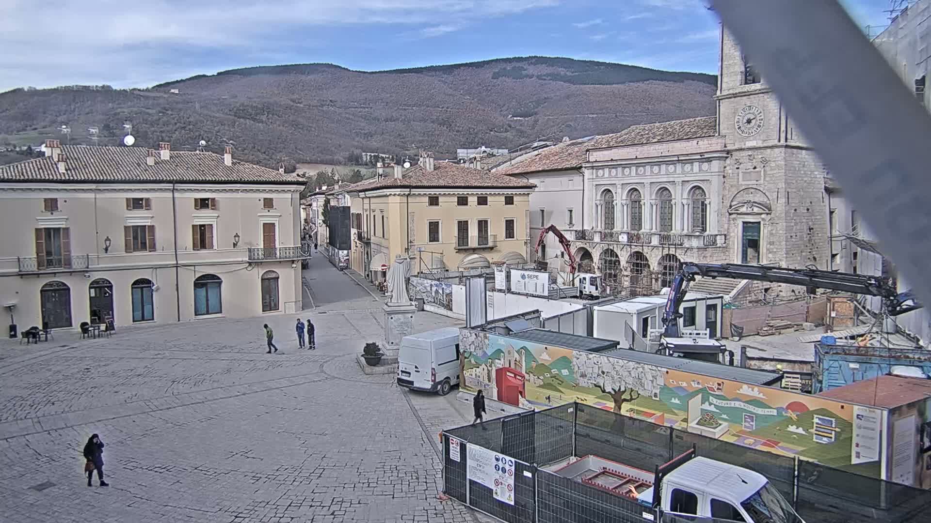 A town square under a partly cloudy sky, with construction equipment and temporary structures in the center, is surrounded by historical buildings, and a few pedestrians are visible.