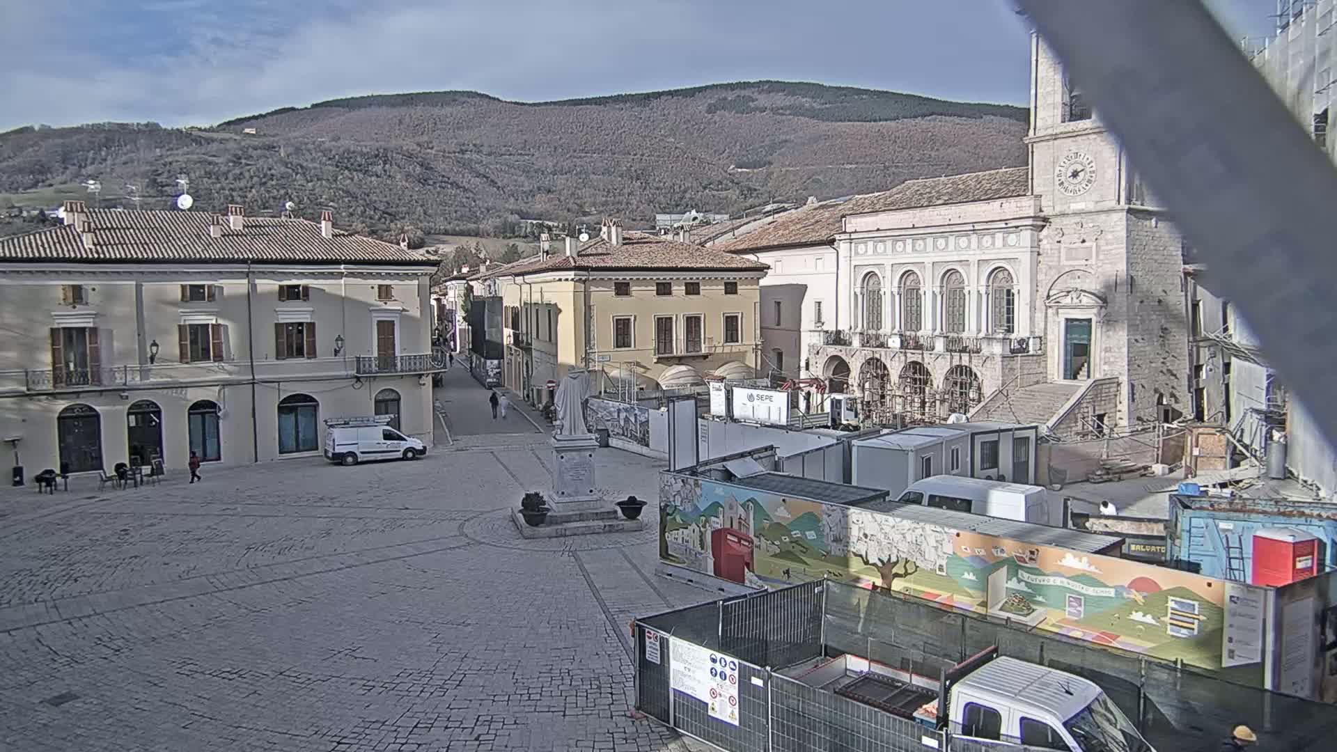 A town square with several buildings, some under construction, is situated against a backdrop of mountains under a partly sunny sky.