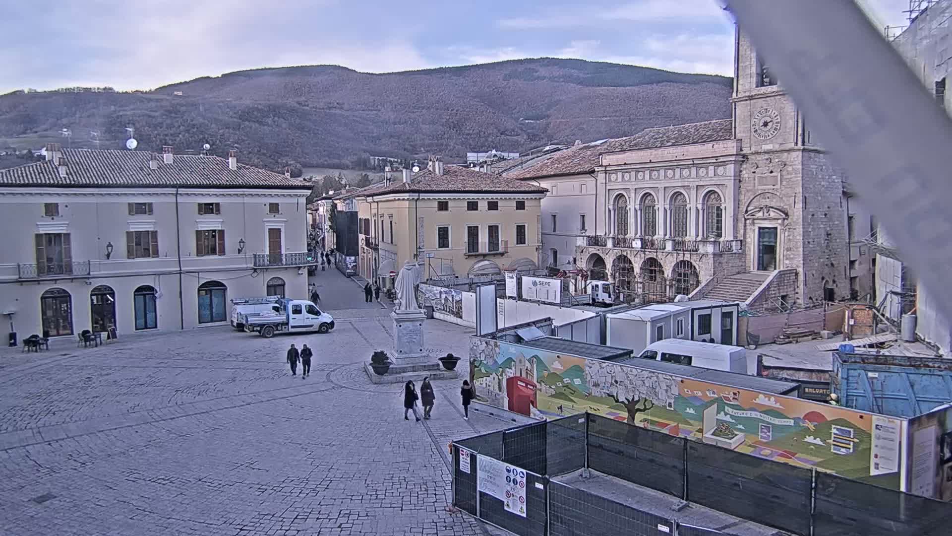 A mostly empty cobblestone town square is under construction, with buildings surrounding it, under a cloudy sky and mountains in the background.