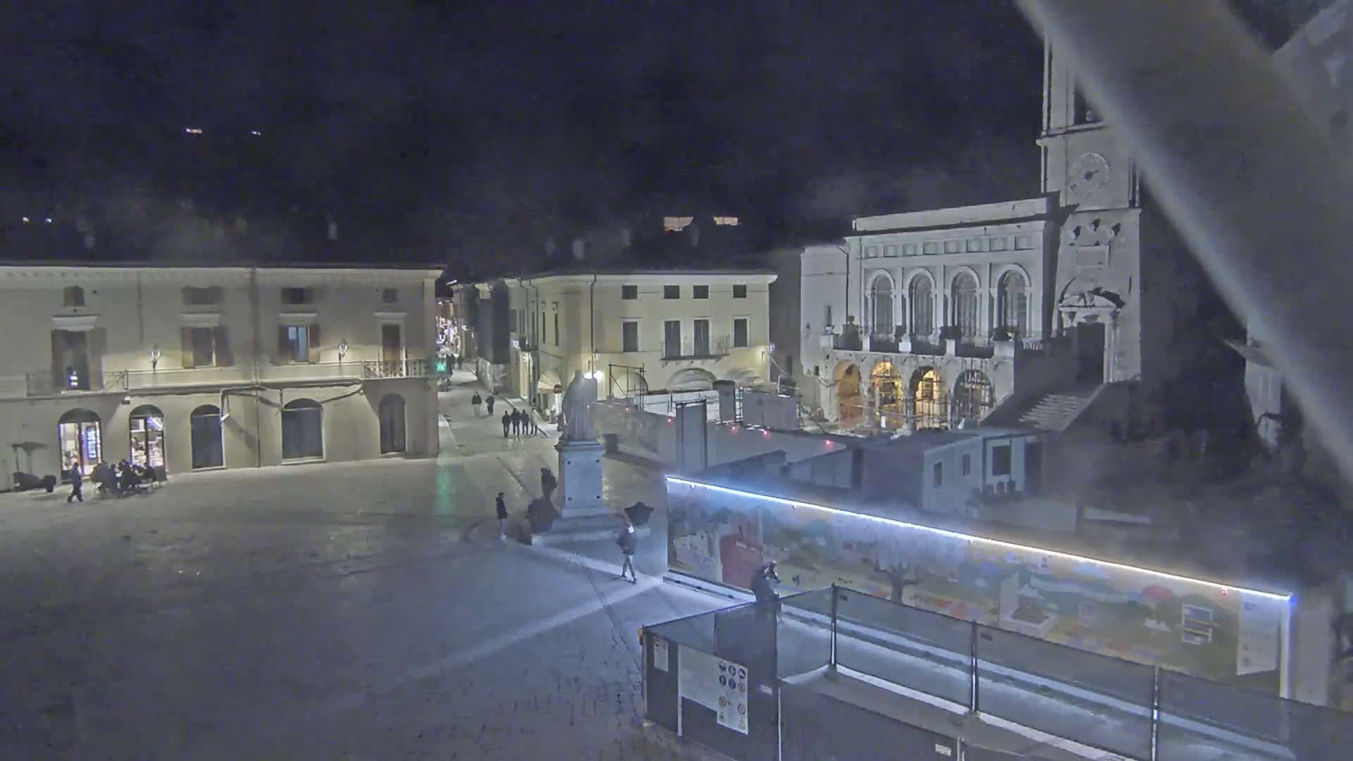 A snow-covered town square at night is surrounded by buildings, including a large church with a clock tower, under a dark sky.