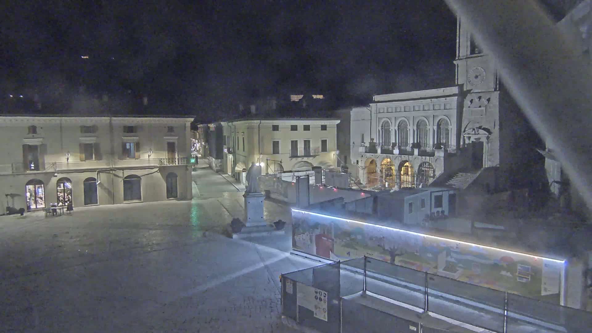 A nighttime view of a town square with several buildings, including a large structure with arched windows, partially obstructed by construction barriers and a painted wall.