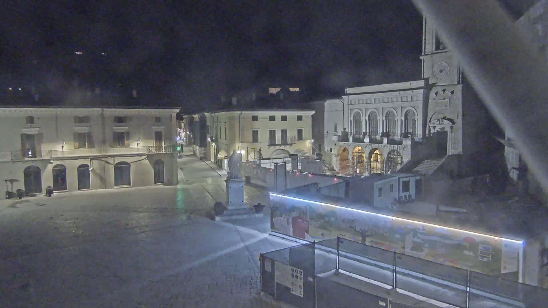 A nighttime view of a town square with several buildings, a statue, and construction barriers, under overcast conditions.