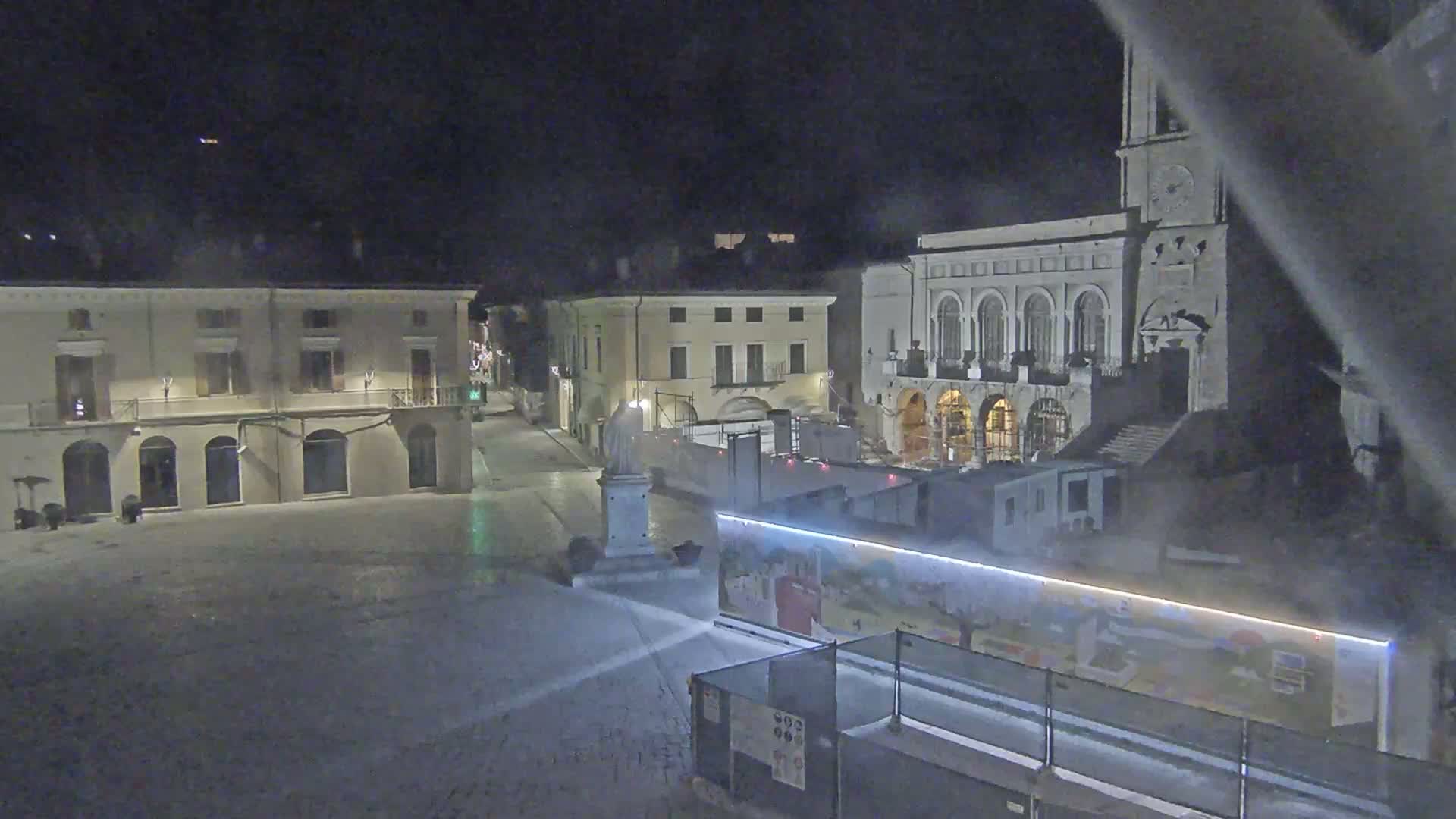 A snow-covered town square at night, featuring several buildings, a statue, and construction barriers with artwork.