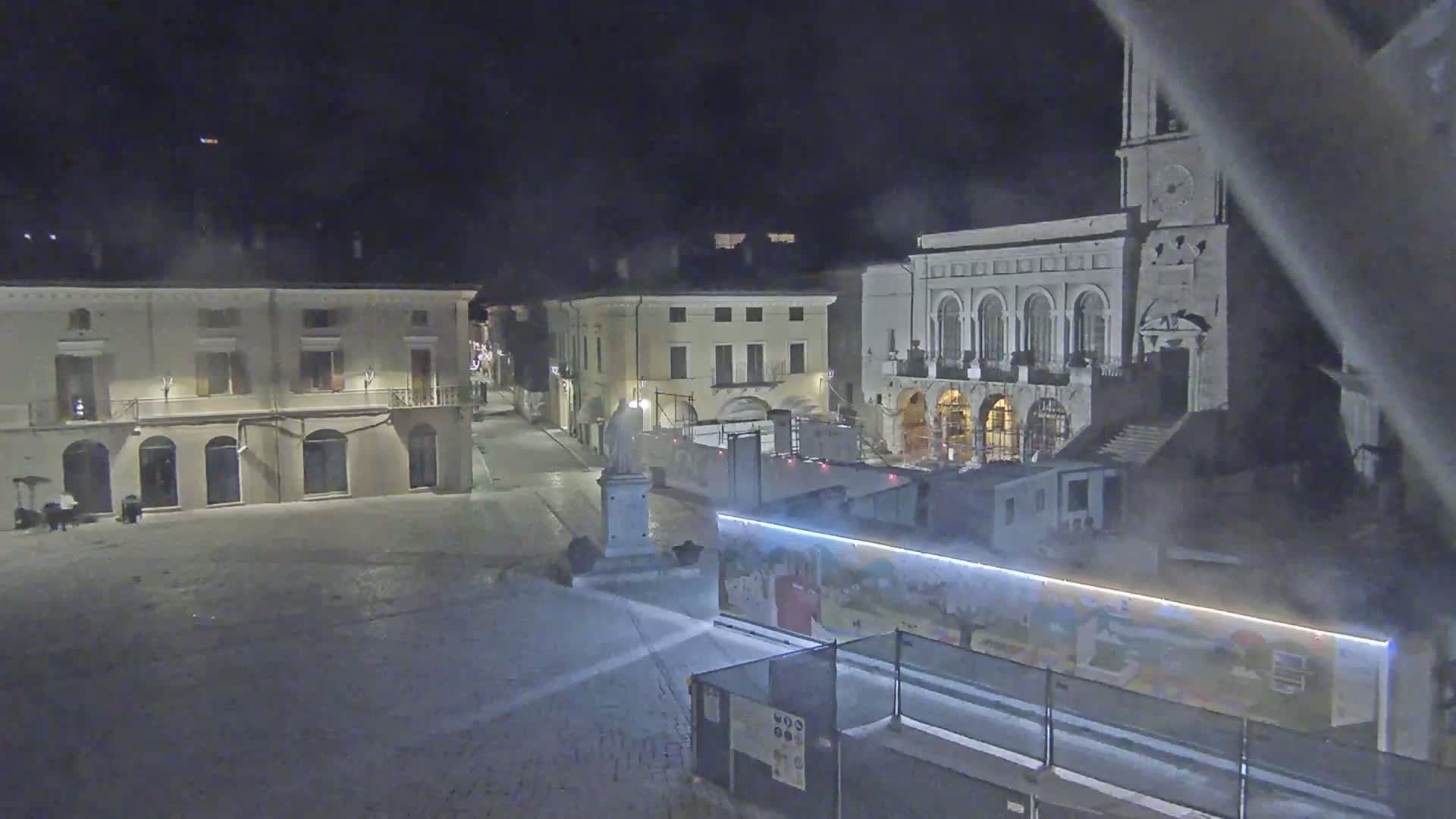 A snow-covered town square at night, with several buildings illuminated, one of which is a large church, and construction barriers in the foreground.