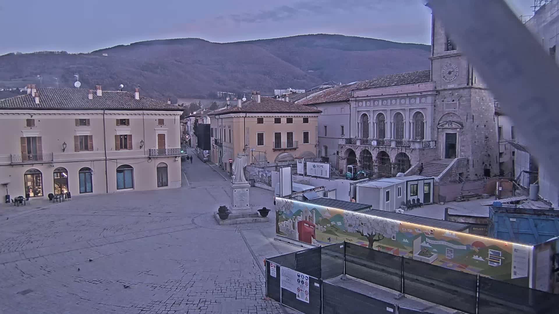 A mostly empty cobblestone town square is under construction, with buildings lining the sides and mountains visible in the background under a dusky sky.