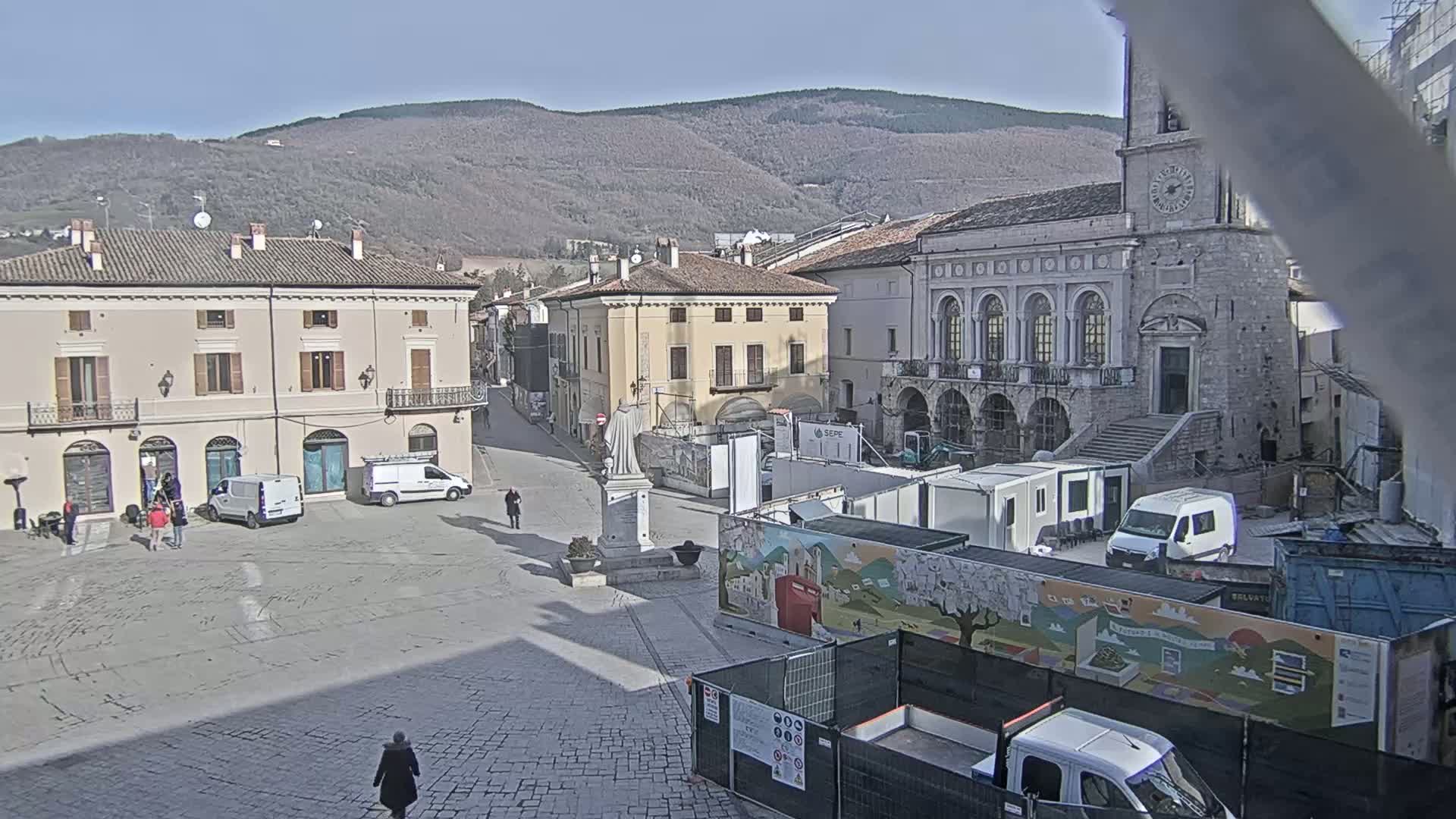 A town square with several buildings, some construction equipment, and a few pedestrians, under a sunny sky with mountains in the background.