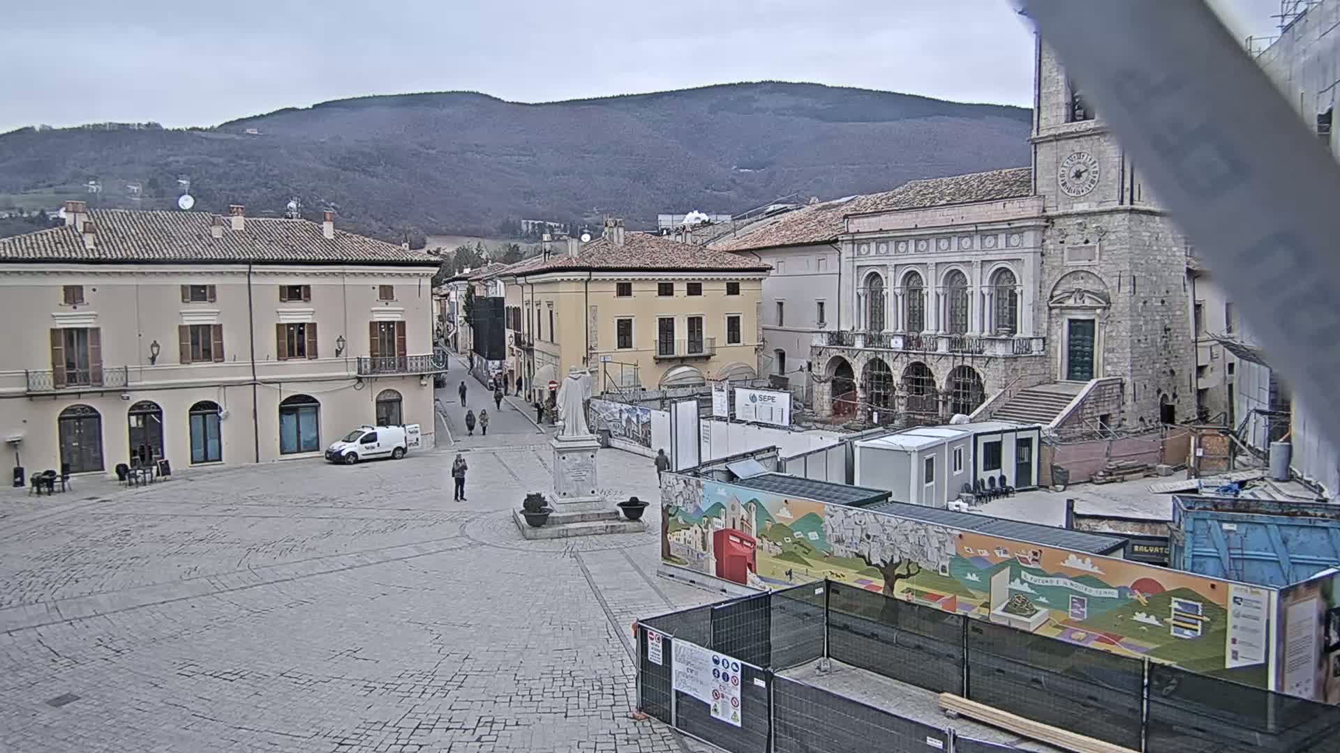 A mostly empty cobblestone town square is under construction, with buildings and a hill visible in the background under an overcast sky.