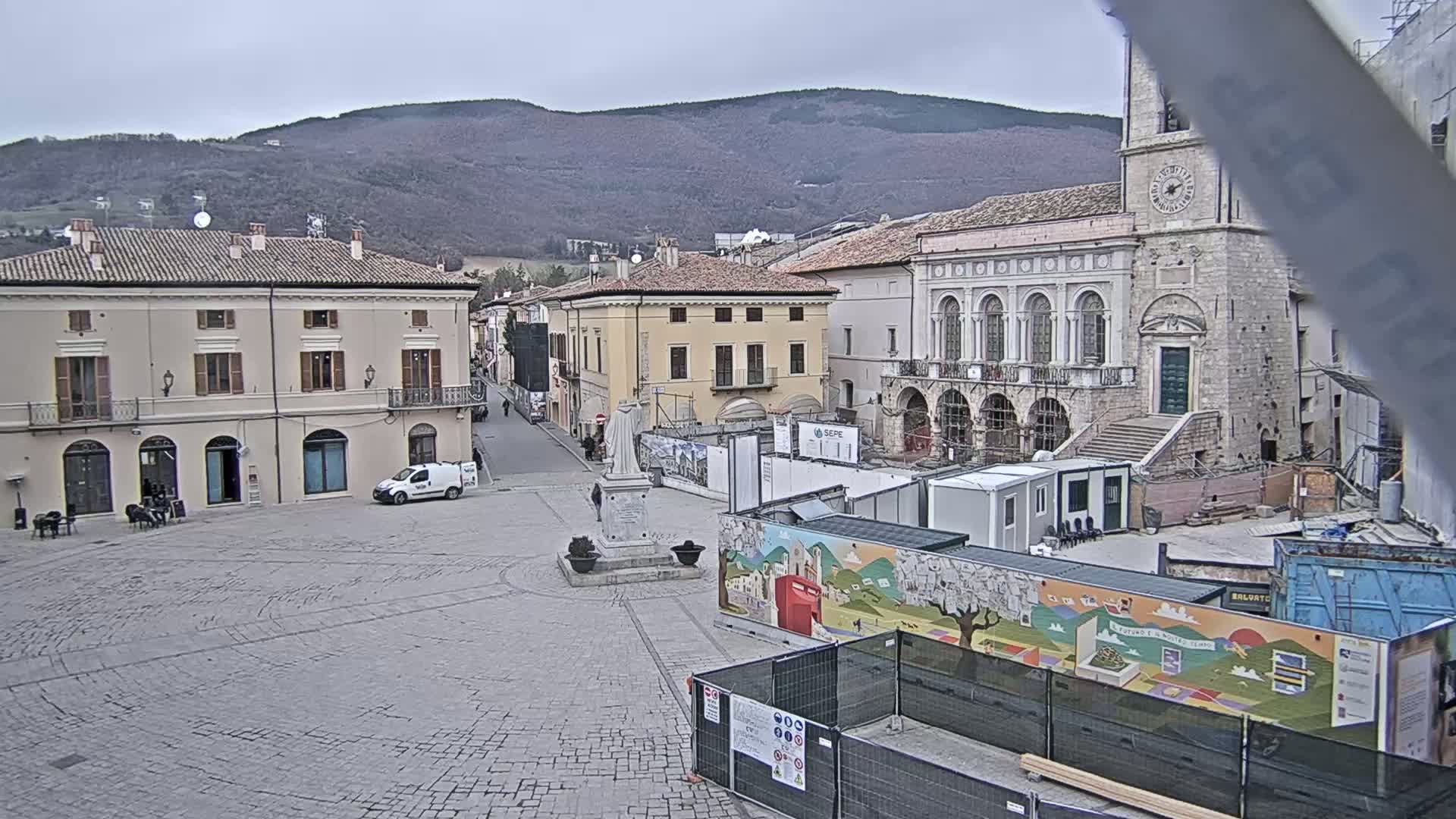 A town square with stone paving, surrounded by buildings, under a cloudy sky, with construction underway in the square's center.