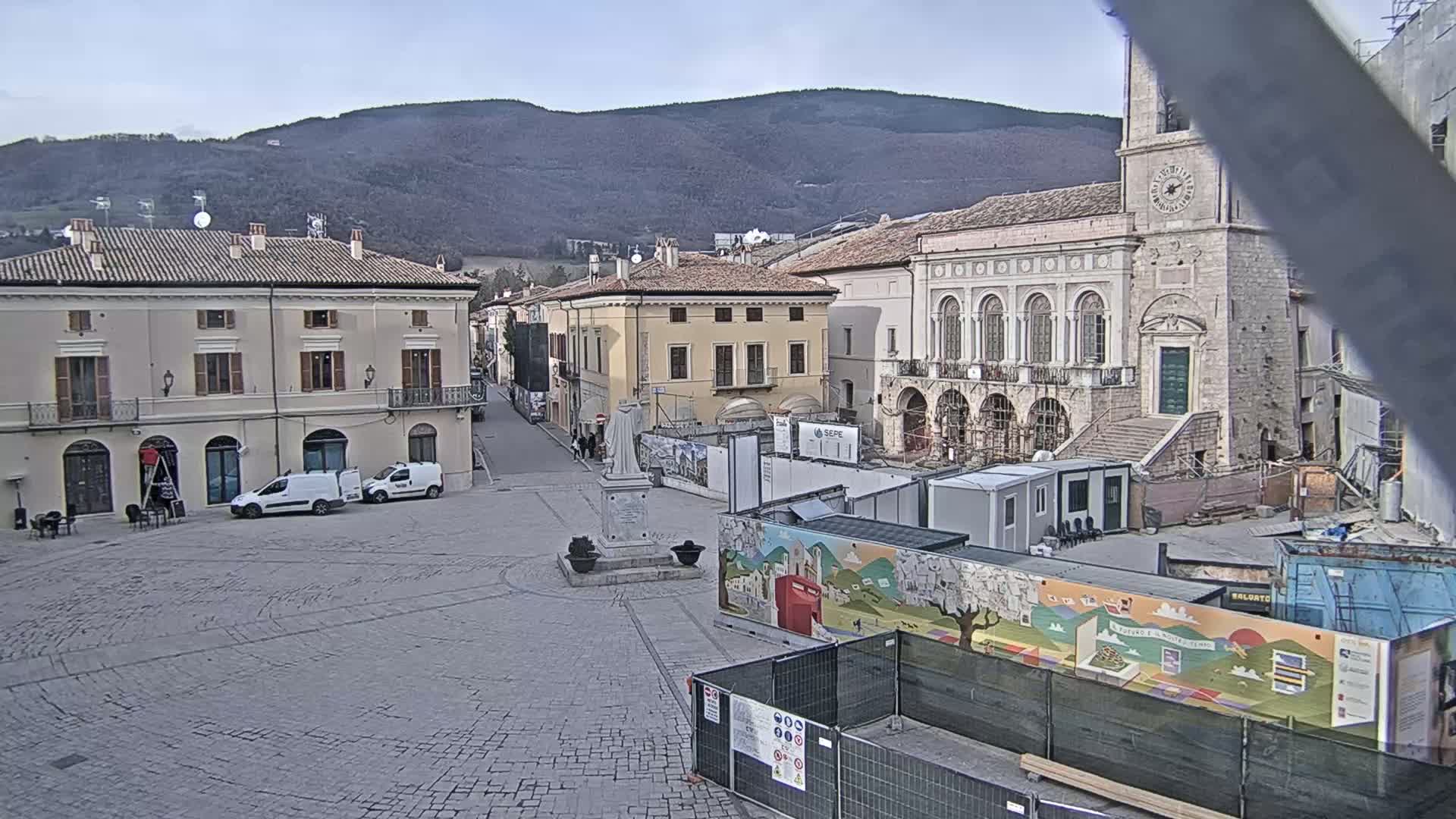 A mostly empty cobblestone town square, under a cloudy sky, is partially surrounded by historic buildings undergoing construction, with mountains visible in the background.