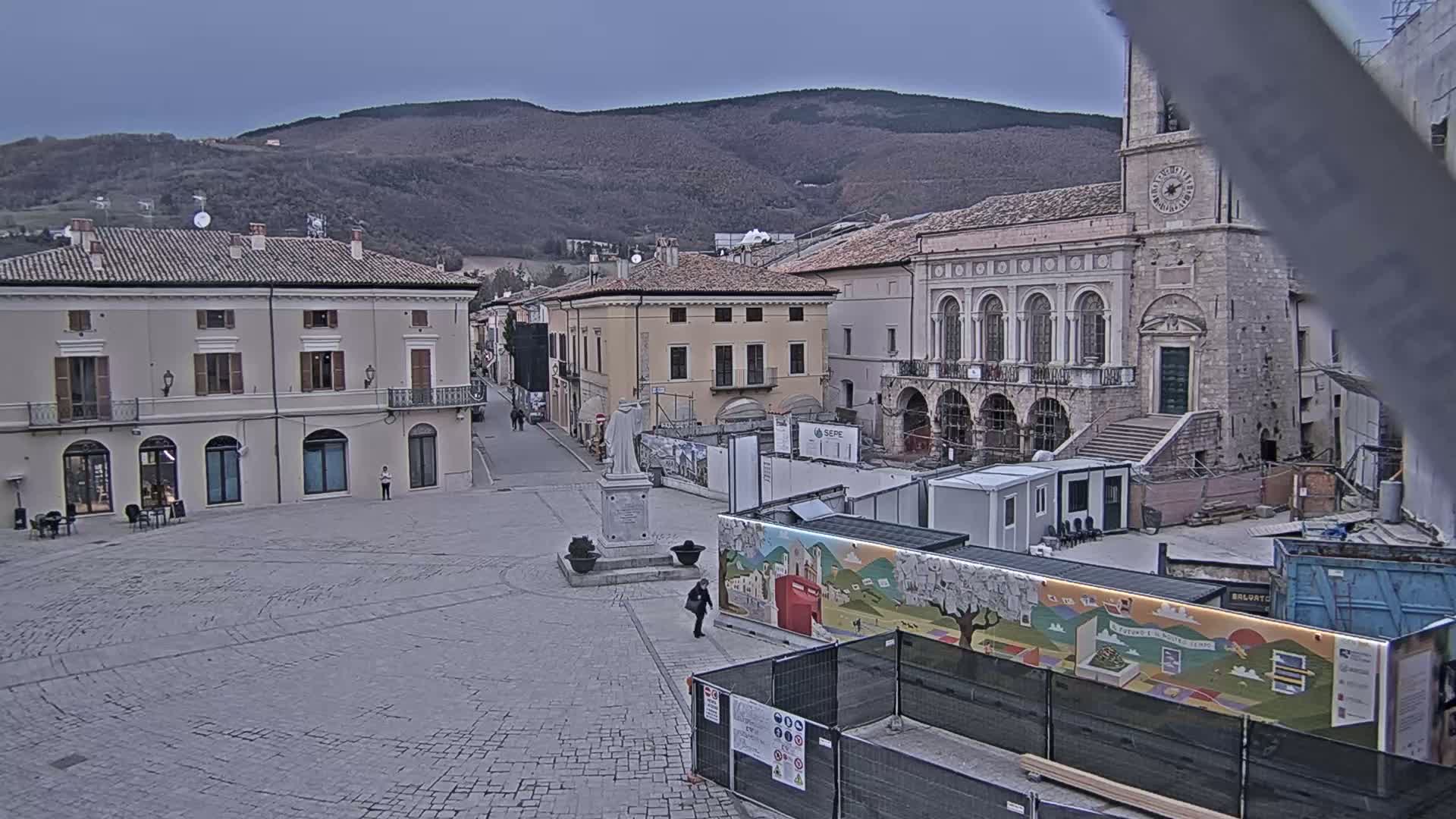 A town square with several buildings, a statue, and construction underway, all under an overcast sky with mountains in the background.