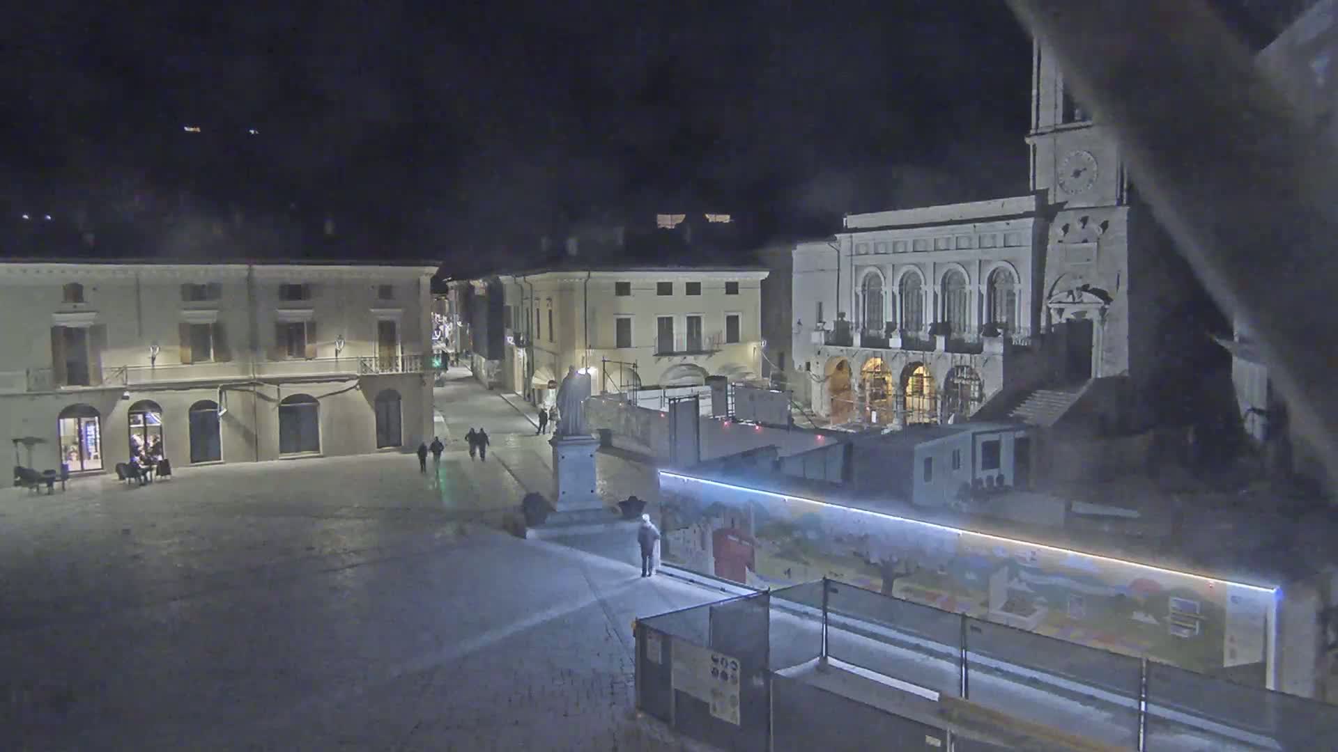 A nighttime view of a town square with several buildings, a statue, and a few pedestrians, under dark and possibly cloudy skies.