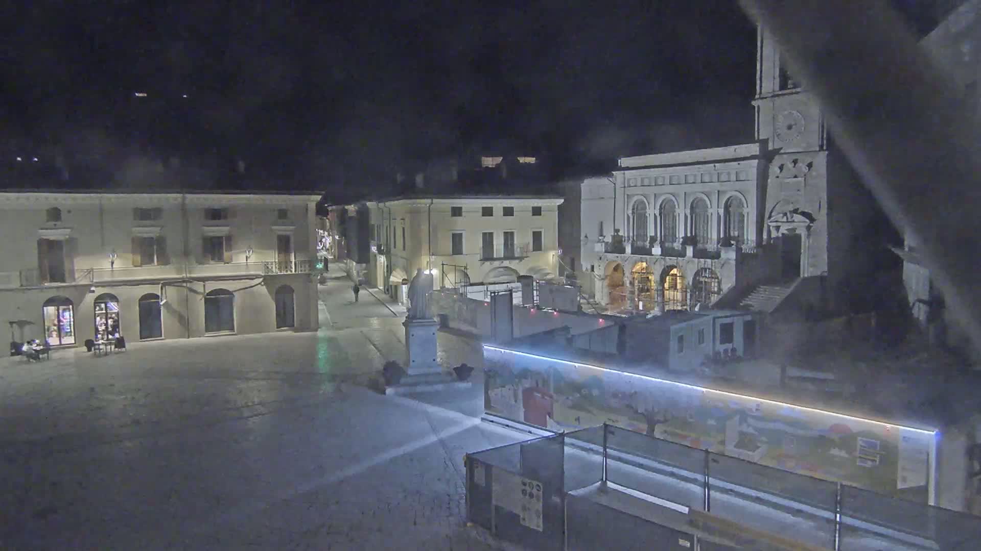 A nighttime view of a town square with several buildings, one of which has a clock tower, under a dark sky.