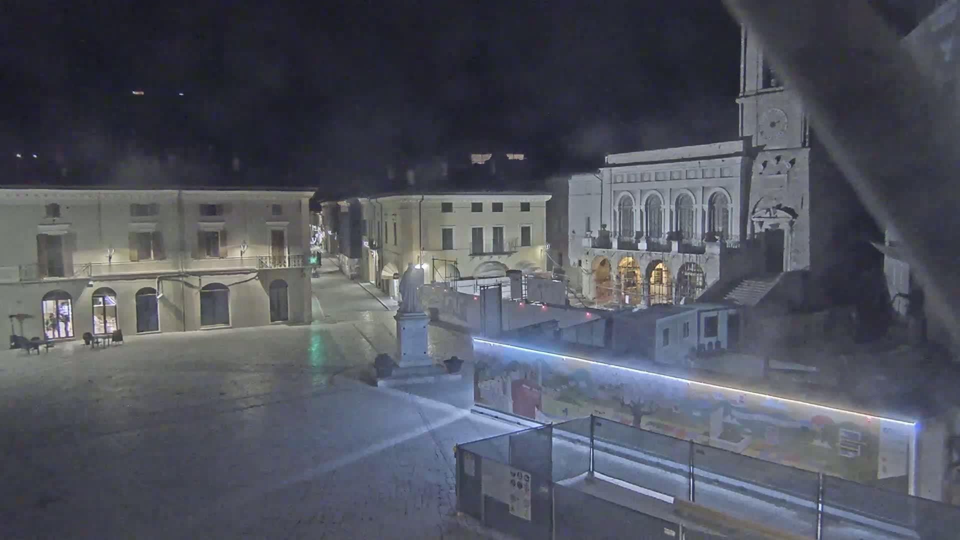 A nighttime view of a town square, under hazy conditions, shows several buildings, including a large church with a clock tower, and construction barriers surrounding a mural.