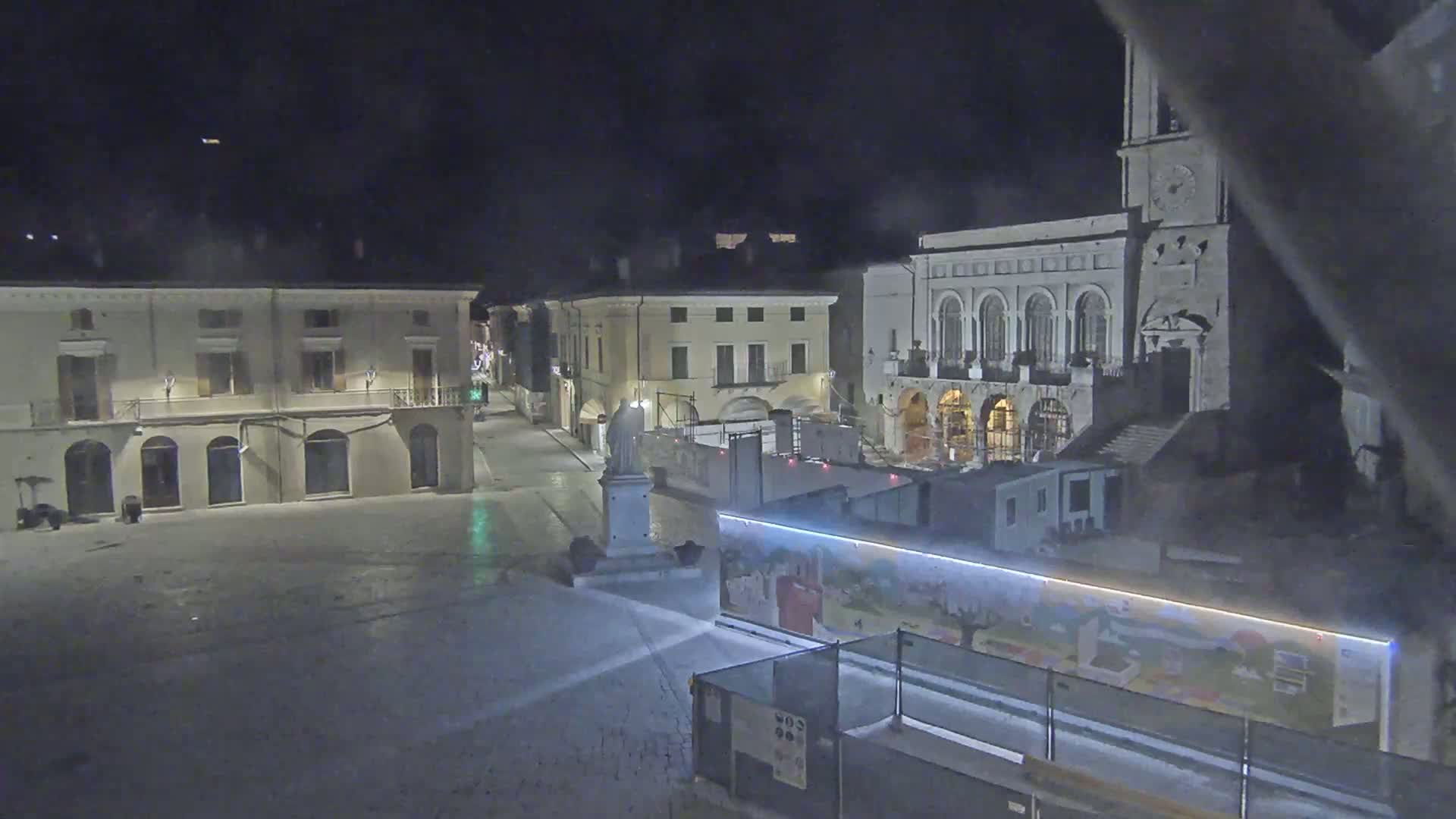 A nighttime, empty town square is shown, featuring several large buildings, a statue, and construction barriers with painted murals.