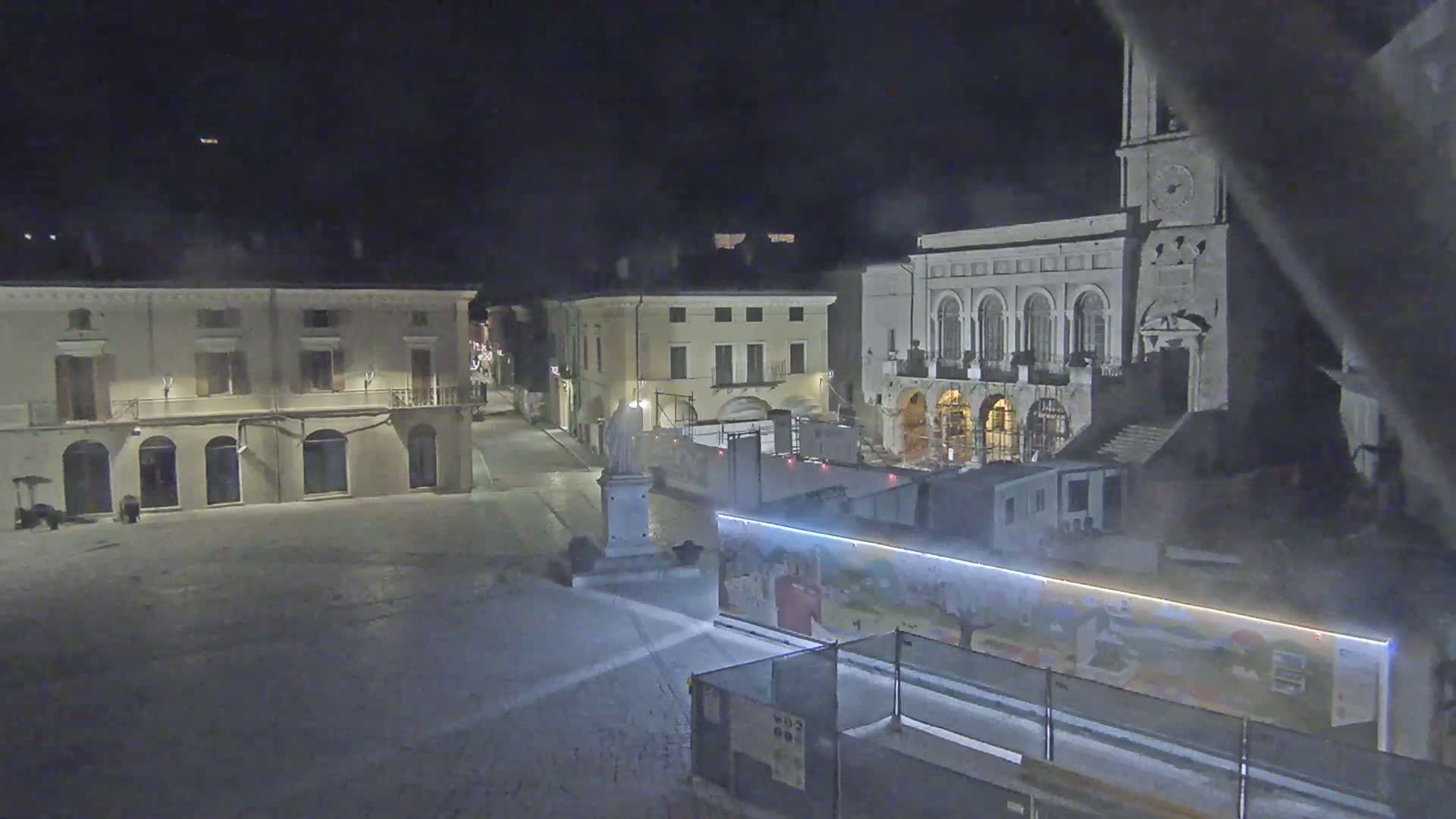 A nighttime view of a town square with several buildings, one of which is a large building with arched windows and a clock tower, partially obscured by construction barriers and mist.