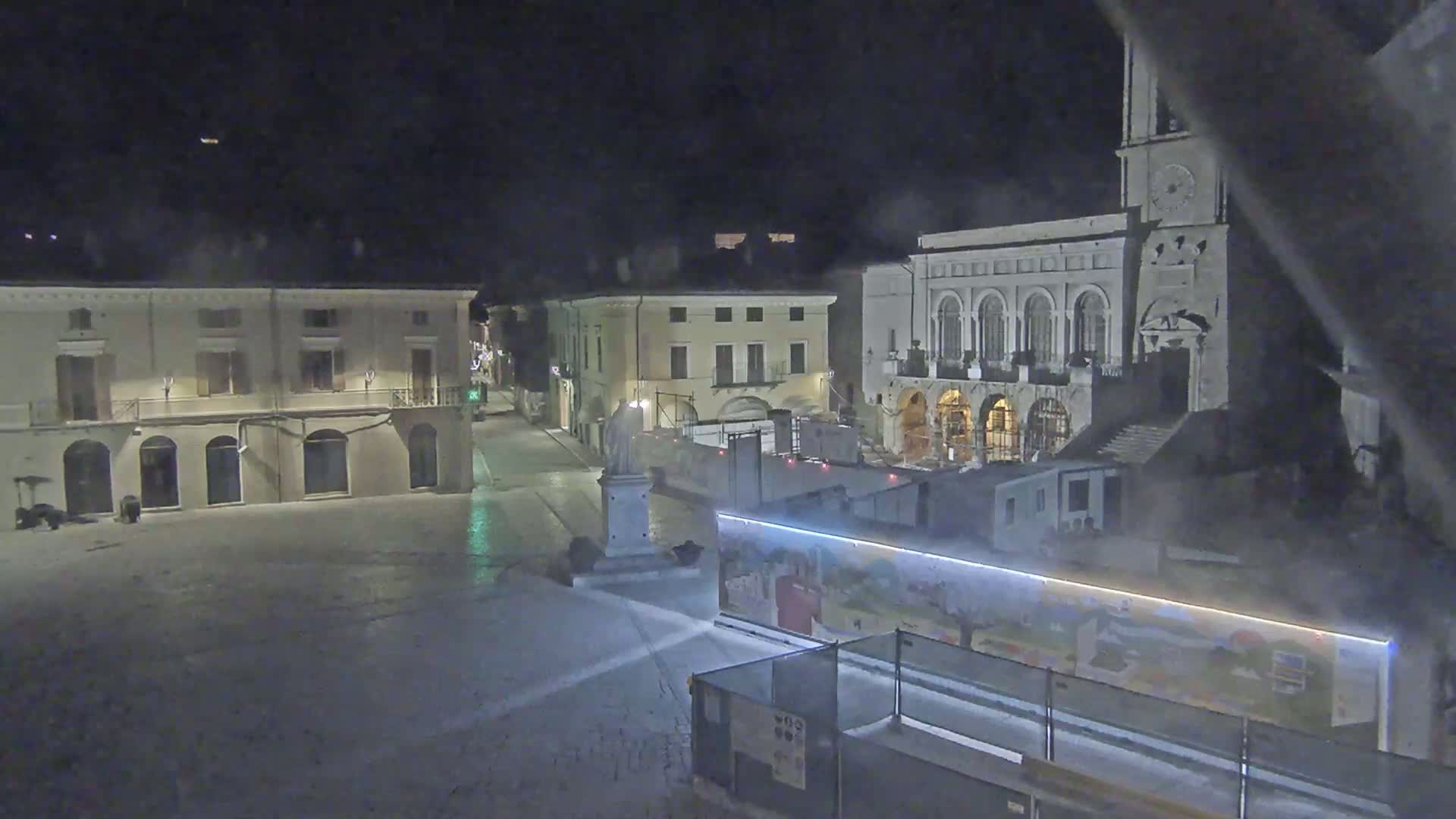 A nighttime view of a town square shows several buildings, a statue, and construction barriers under a dark, possibly foggy sky.