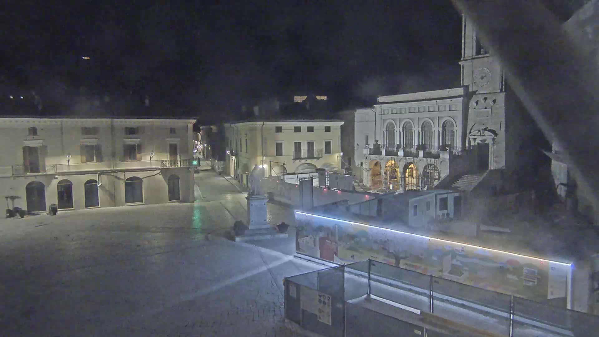 A nighttime view of a town square with several buildings, one of which is a large building with arched windows and a clock tower, partially obscured by construction barriers and fog or mist.