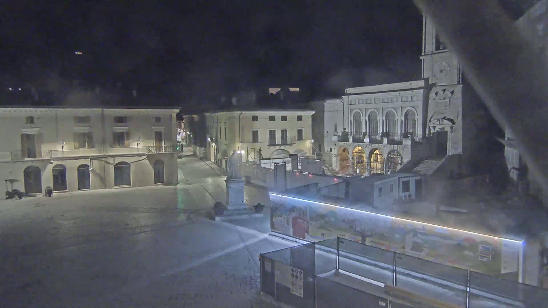 A nighttime view of a town square with several buildings, a statue, and construction barriers, under cloudy conditions.
