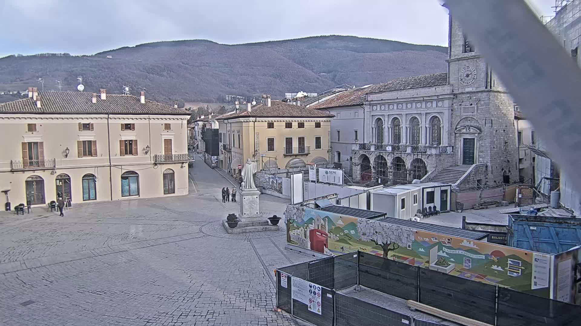 A town square with stone buildings and a statue, under a cloudy sky, is partially under construction.