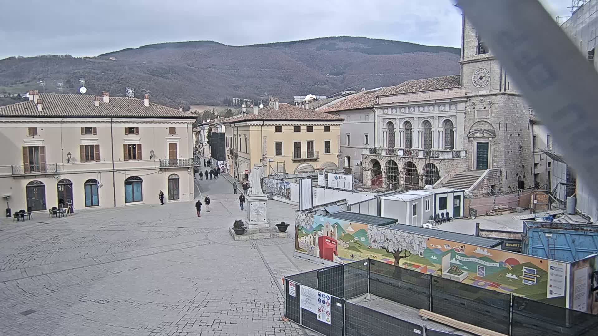 A mostly empty cobblestone town square with several buildings, a statue, and construction underway under an overcast sky, backed by a range of distant, bare-branched hills.