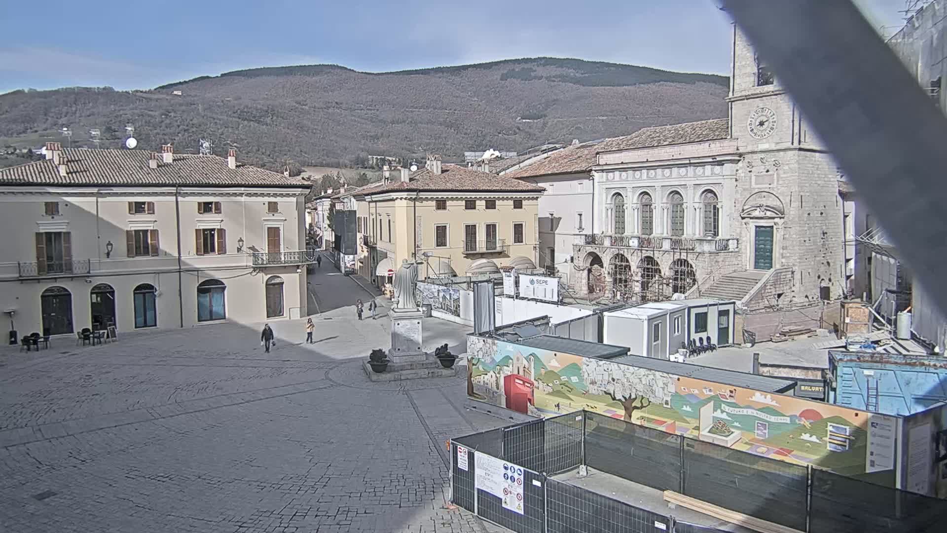 A town square with several buildings, under construction, is surrounded by mountains under a clear, sunny sky.