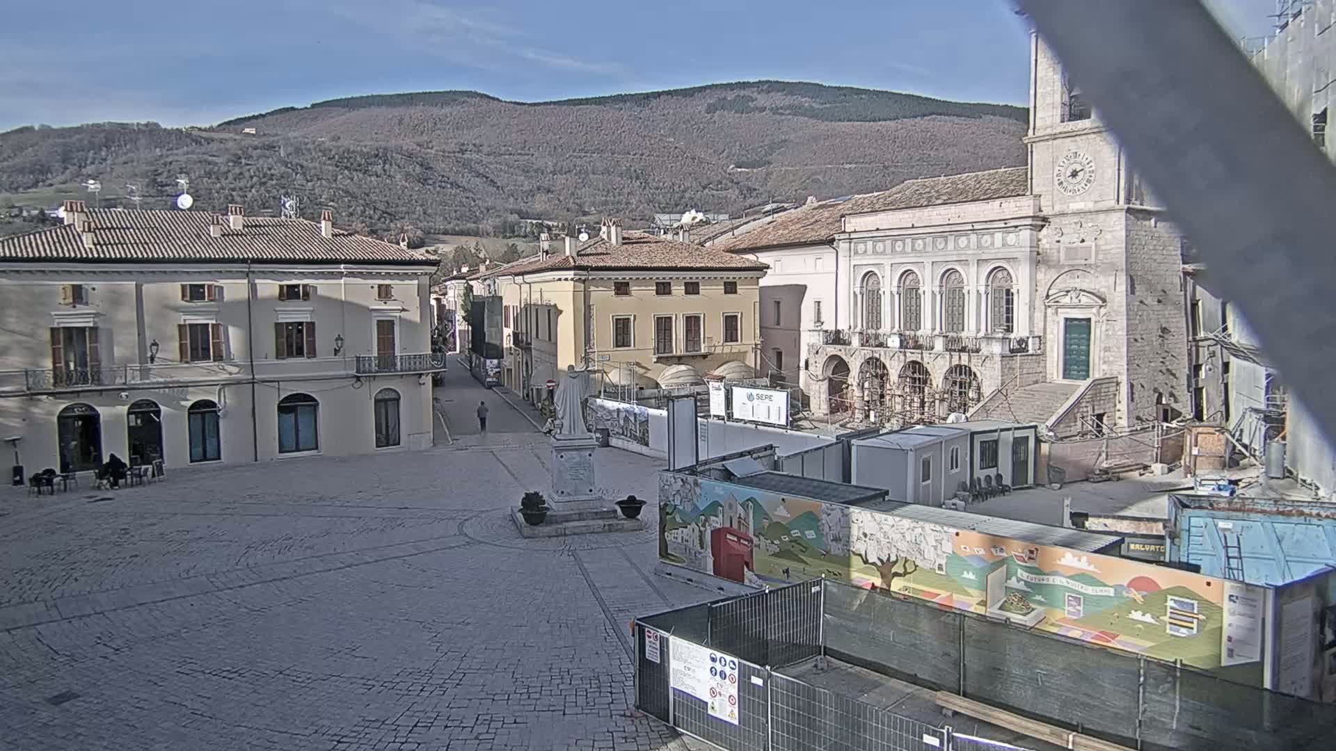 A town square under a sunny sky is partially blocked off by construction barriers and temporary buildings, with mountains visible in the background.