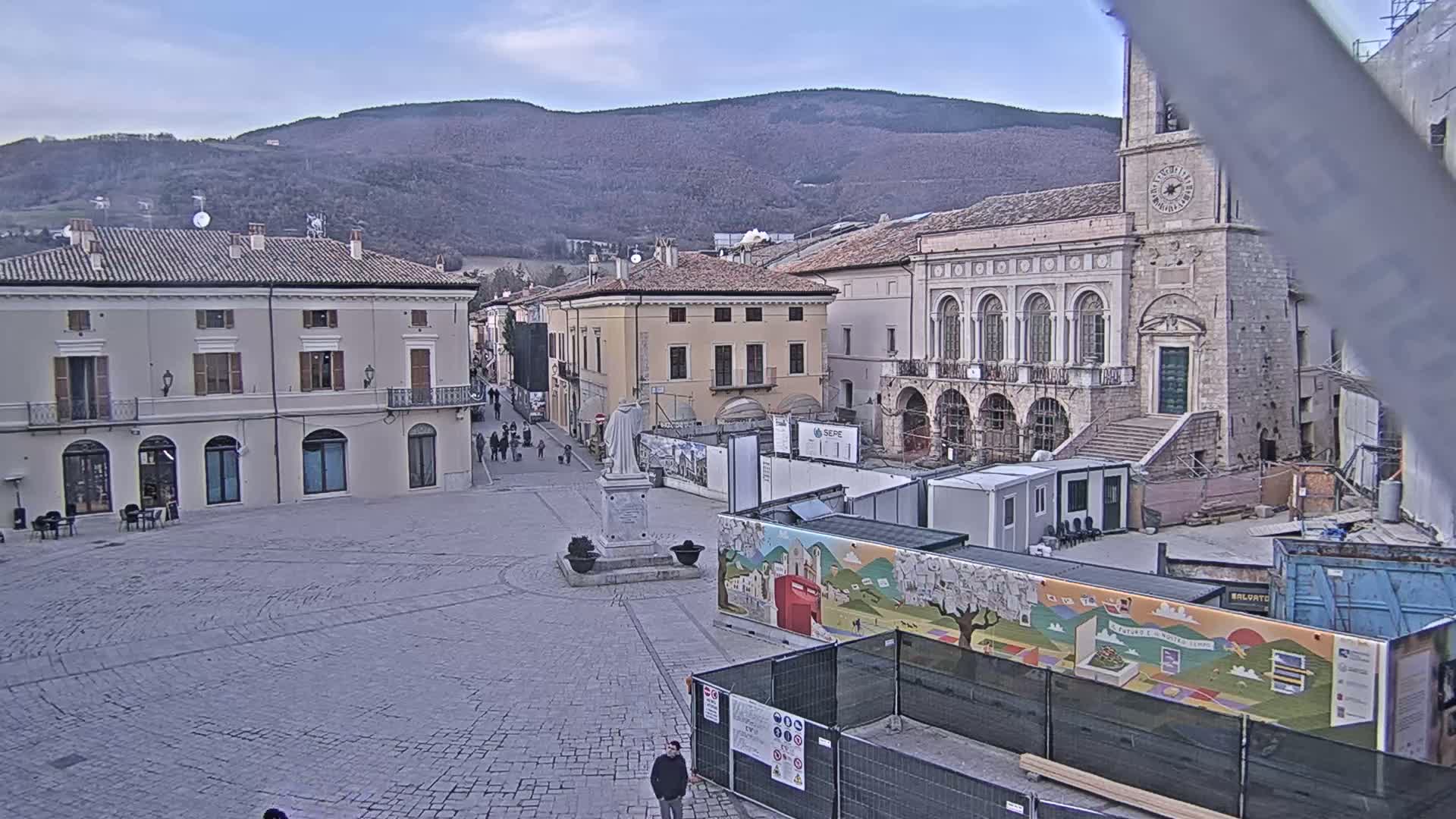 A town square with stone buildings, a statue, and construction barriers under a partly cloudy sky, with mountains visible in the background.