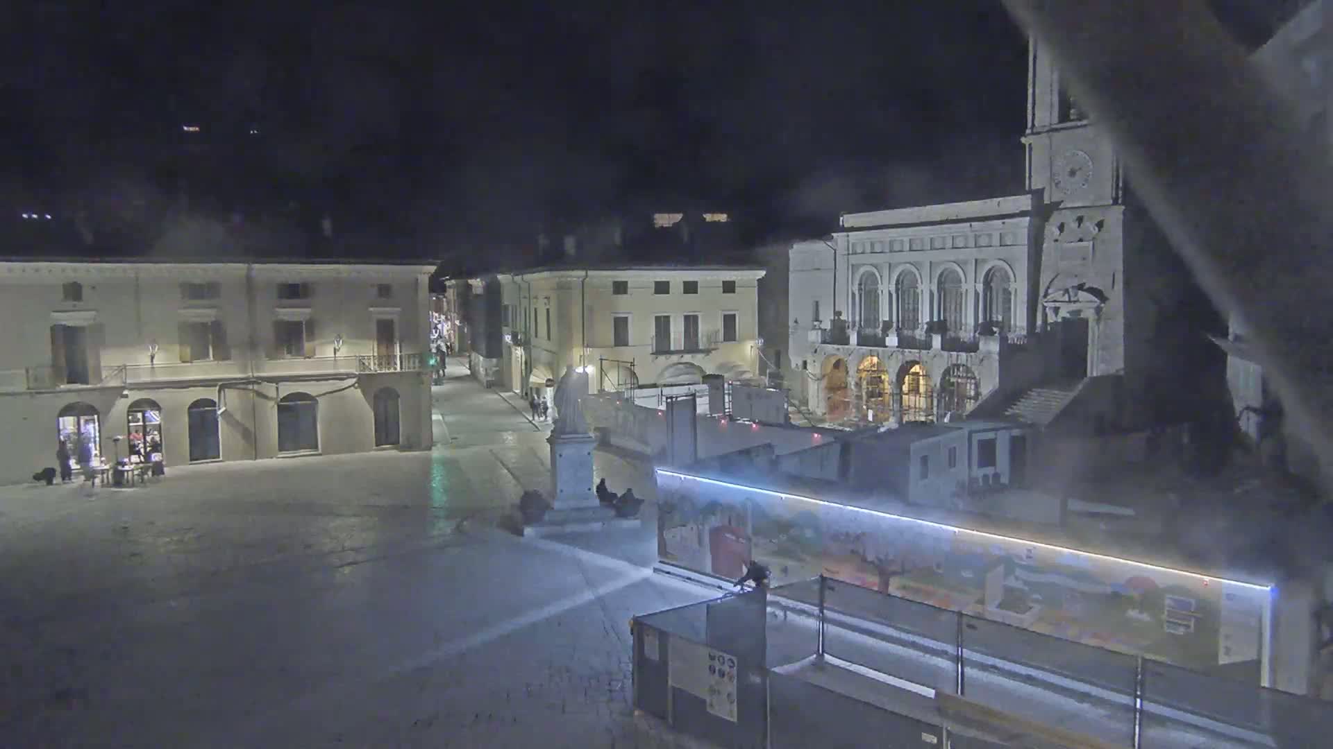 A snow-covered town square at night is surrounded by buildings, including a large church with a clock tower, and construction barriers are set up near a painted wall.