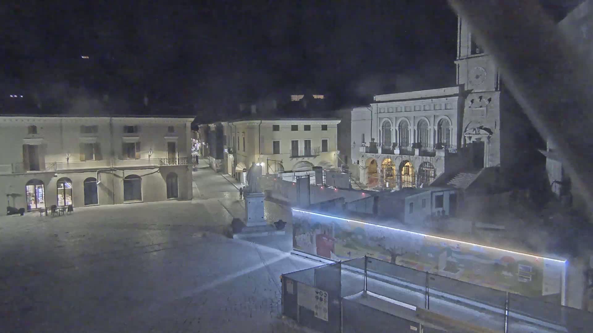 A snow-covered town square at night, featuring several buildings, one of which is a large church with a clock tower, and construction barriers with artwork displayed.